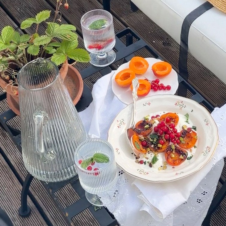 Glaskrug und zwei Gläser mit Minzwasser auf Tisch mit Tellern mit Aprikosen und roten Johannisbeeren