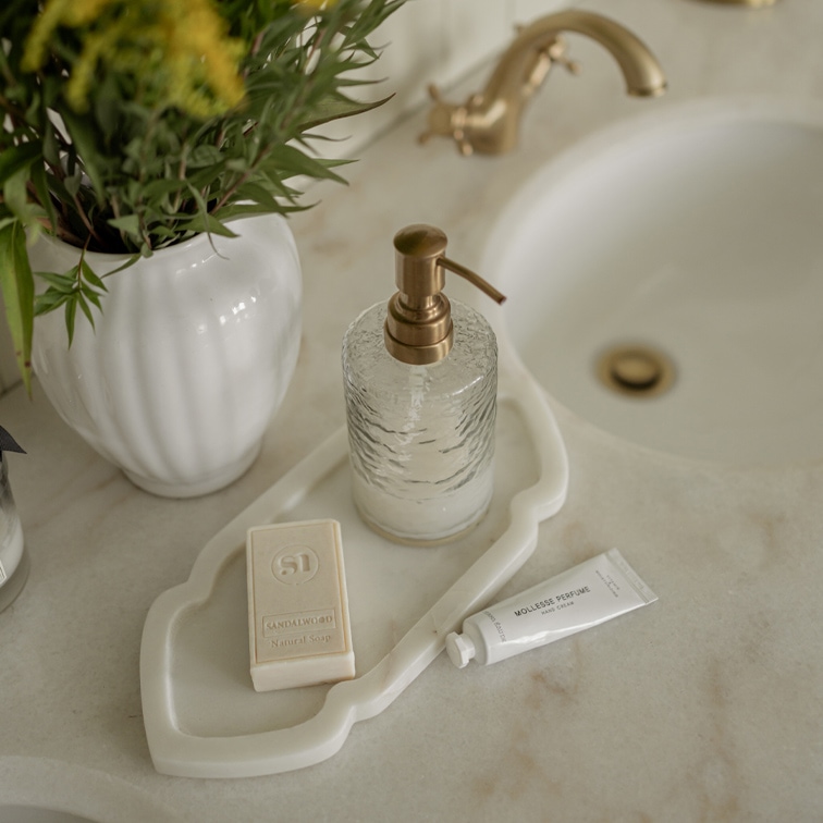 Glas-Seifenspender mit Messingpumpe auf weißem Tablett neben Seife und Handcreme auf Marmorwaschbecken