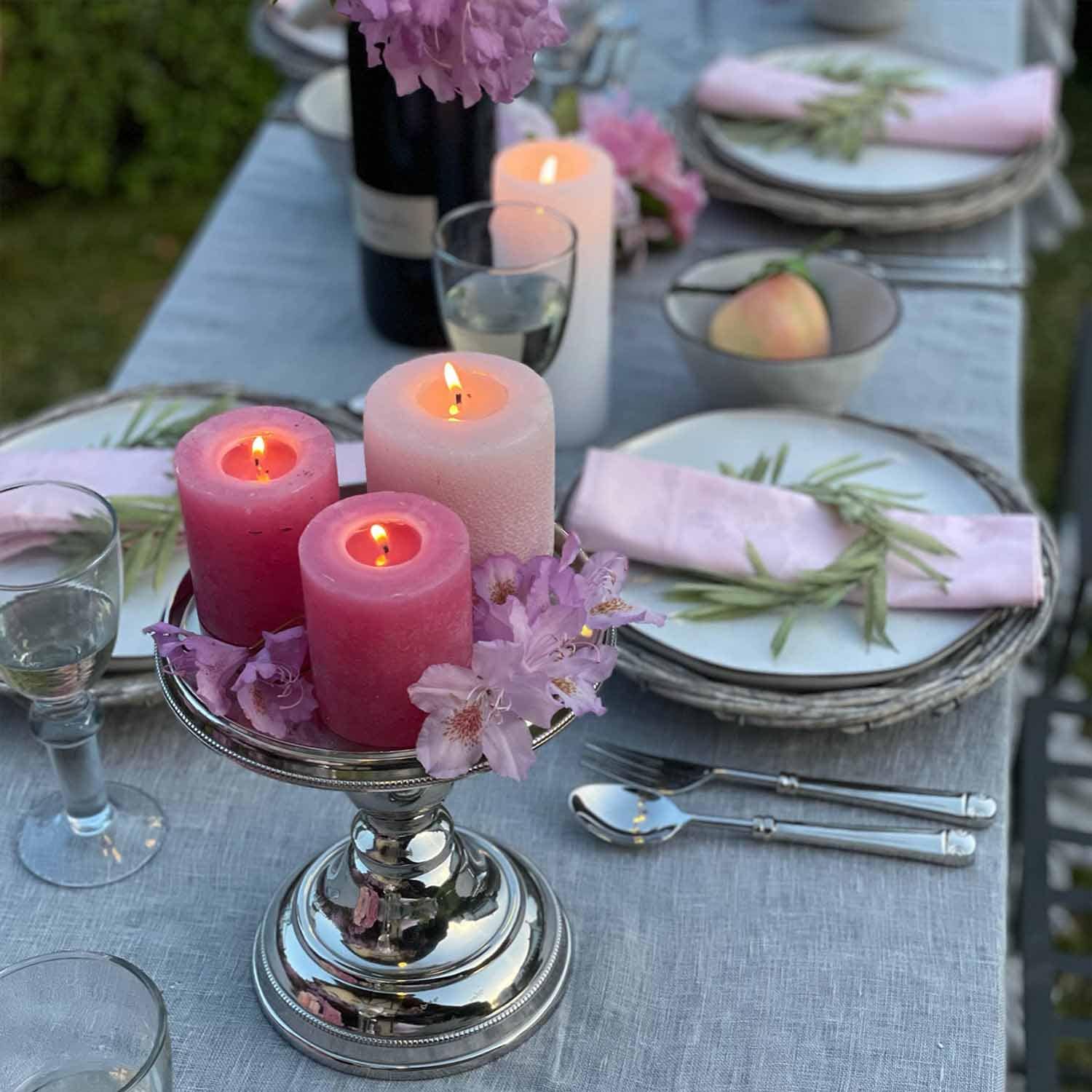 Silberner Kerzenhalter mit drei brennenden rosa und weißen Kerzen auf grauem Tisch mit Blumen