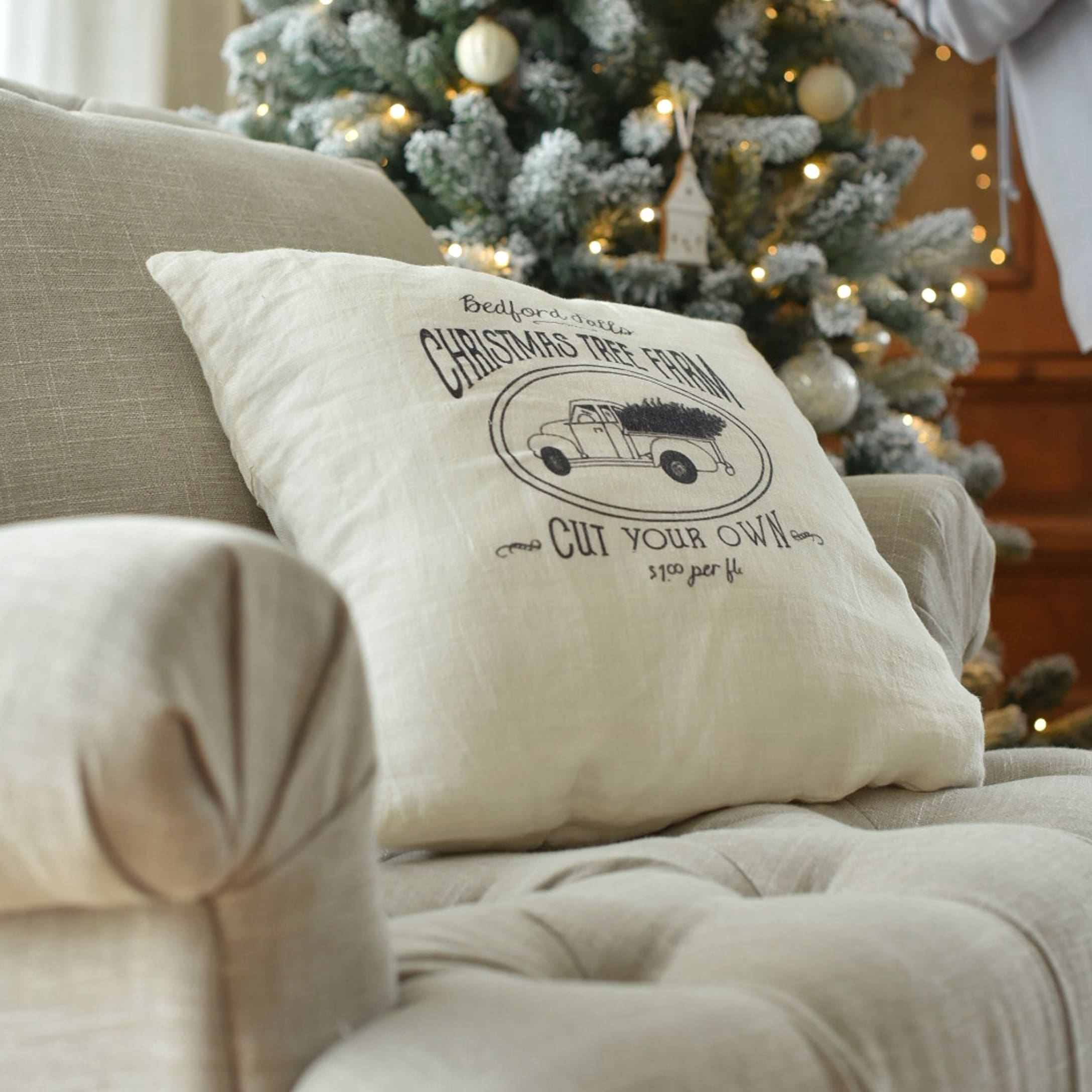 Beiges Leinenkissen mit Aufdruck eines Weihnachtsautos auf Sofa neben festlich beleuchtetem Baum