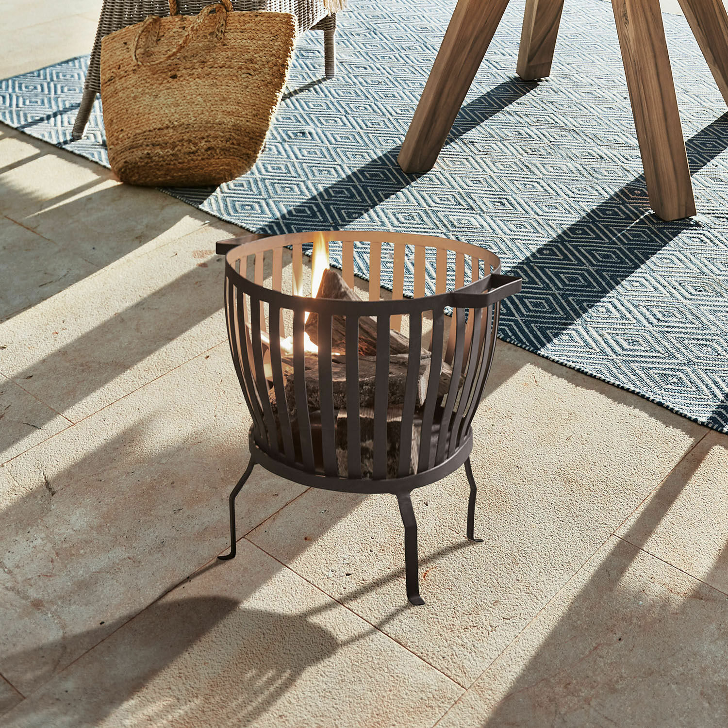 Schwarzer Metall-Feuerkorb mit brennendem Holz auf Terrasse mit blauem Teppich und Korb