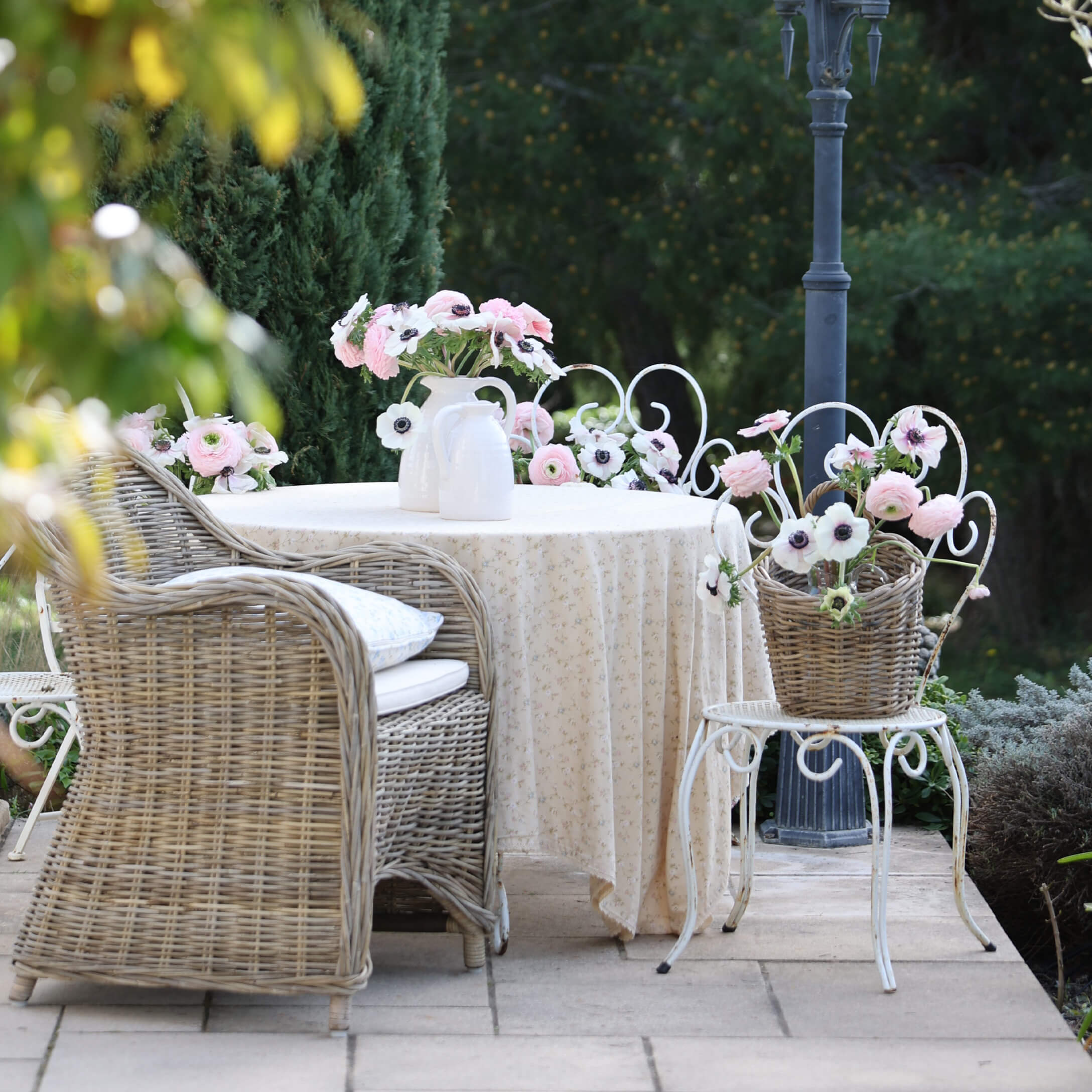 Garten mit Korbsessel, weißem Tisch mit Tischdecke und Blumen in Vasen, romantische Dekoration