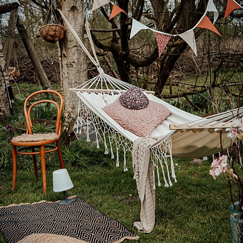 Weißer Stoff-Hängematte mit rosa Kissen und Decke auf grünem Rasen, Holzstuhl und Girlande im Garten