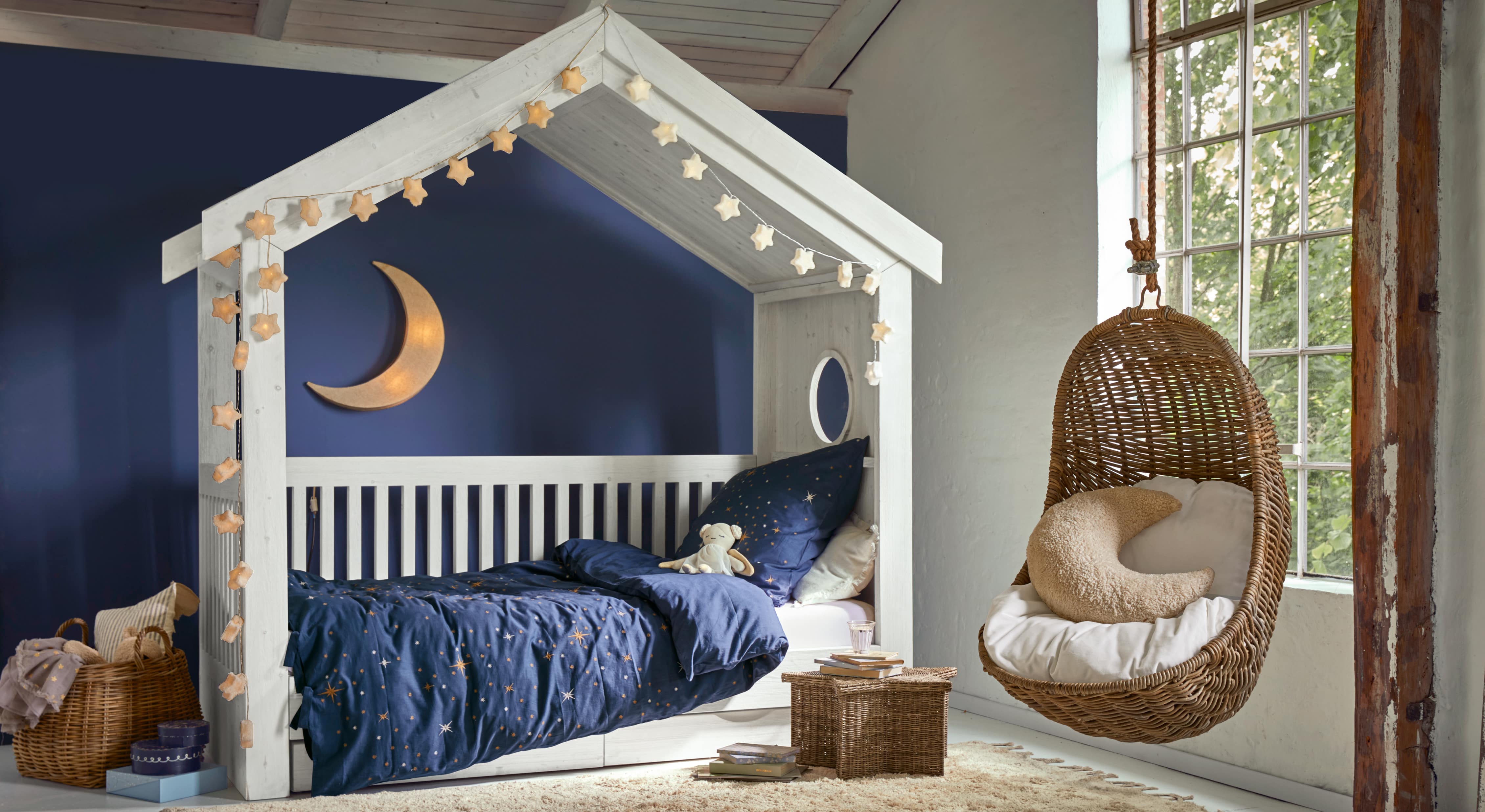 Weißes Holzhaus-Bett mit dunkelblauer Sternenbettwäsche und hängendem Korbsessel im Kinderzimmer