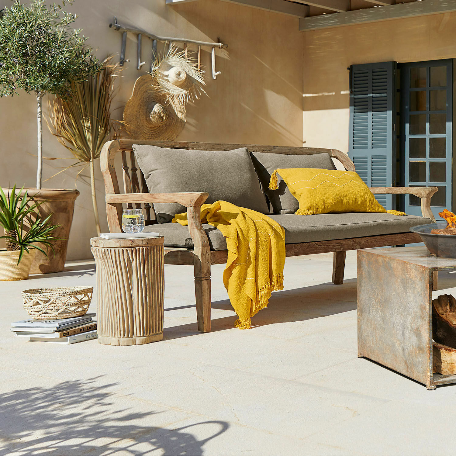 Holzbank mit grauen Kissen und gelber Decke auf Terrasse mit Rattan-Dekorationen und Pflanzen