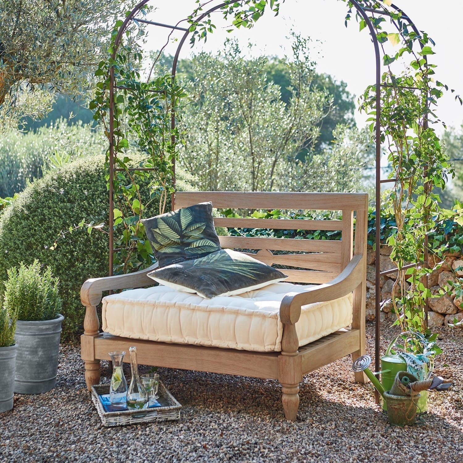 Holz-Gartenbank mit cremefarbenem Polster und grünen Dekokissen auf Kieselstein-Terrasse