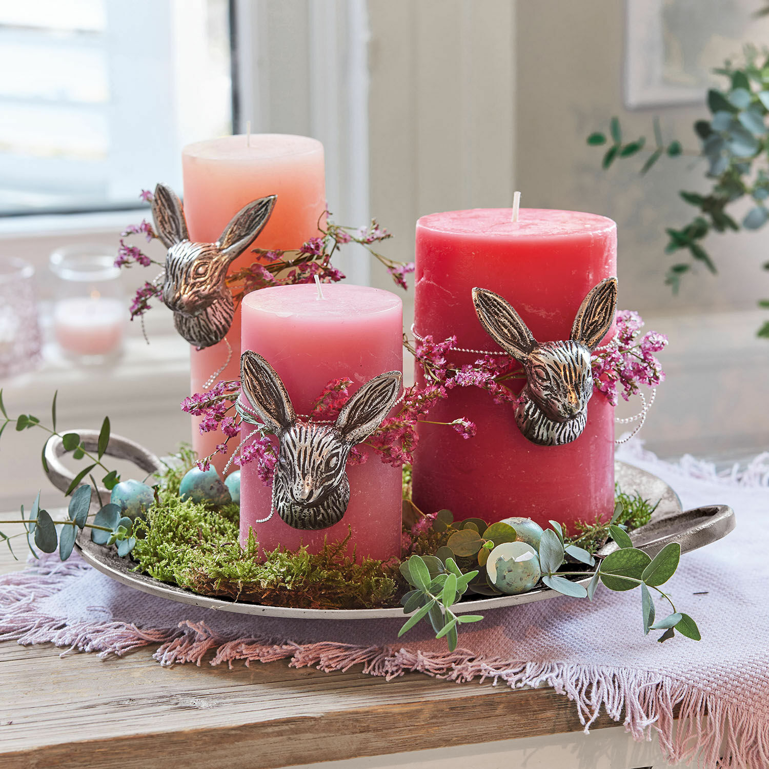 Drei rosa Kerzen mit Metall-Hasenköpfen auf Tablett mit Moos und Osterdekorationen