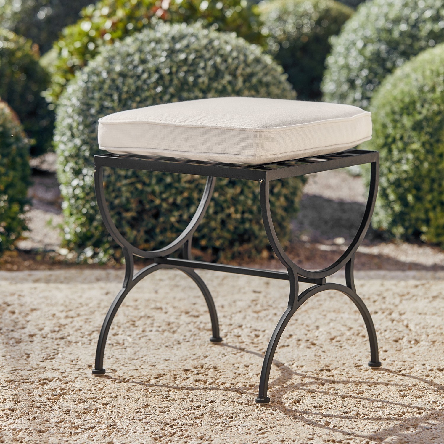 Hocker Terell Metallhocker mit schwarzem Gestell und cremefarbenem Polster auf sandigem Boden im Garten