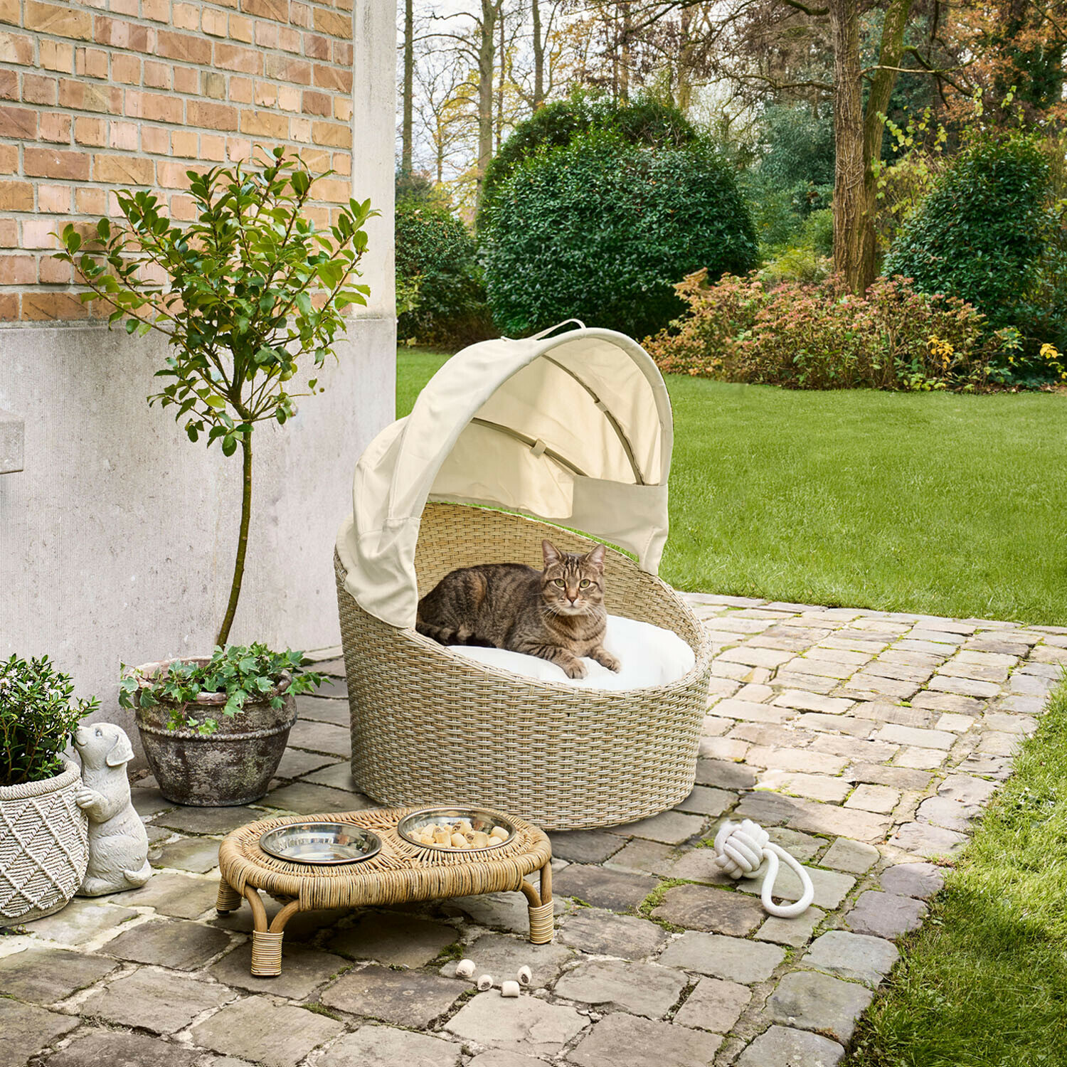In- & Outdoor-Haustierkorb Brinelza Ovales Katzenbett aus beigem Rattan mit weißem Kissen und Dach auf gepflasterter Terrasse im Garten