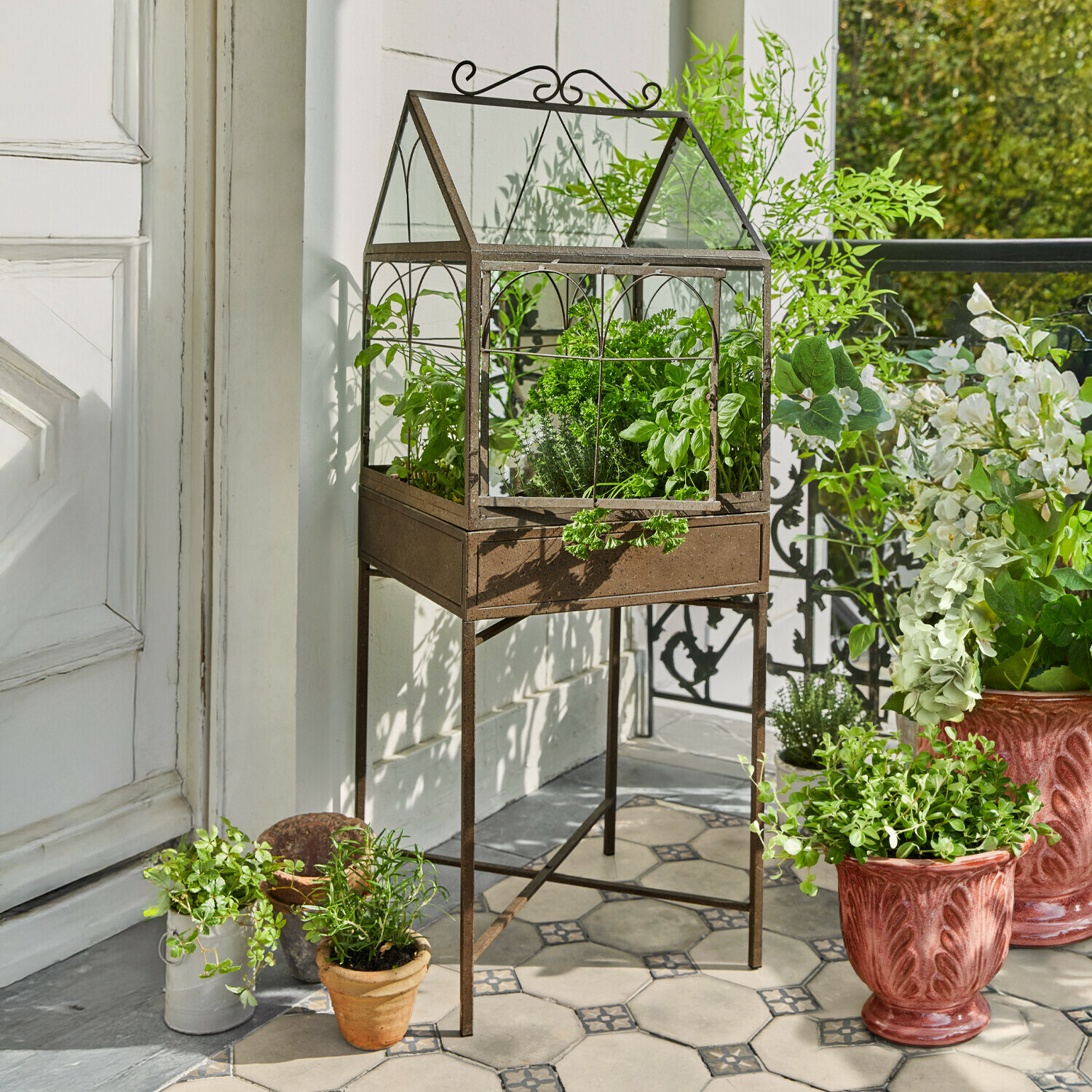 Metall-Gewächshaus mit Rostoptik auf Gestell mit grünen Kräutern auf gefliestem Balkon