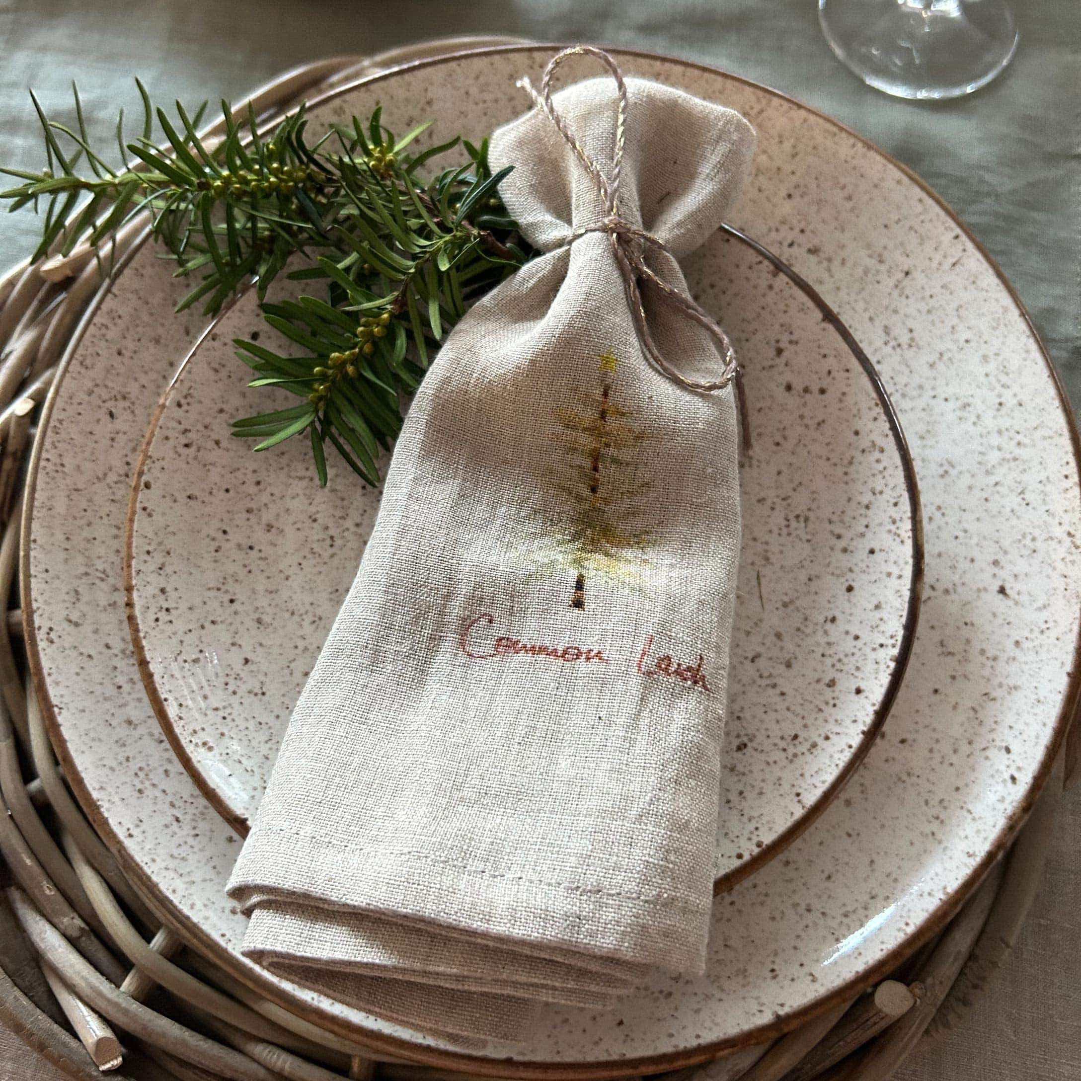 Beige Leinenbeutel mit Tannenbaum-Motiv auf Keramikteller mit grünen Zweigen dekoriert