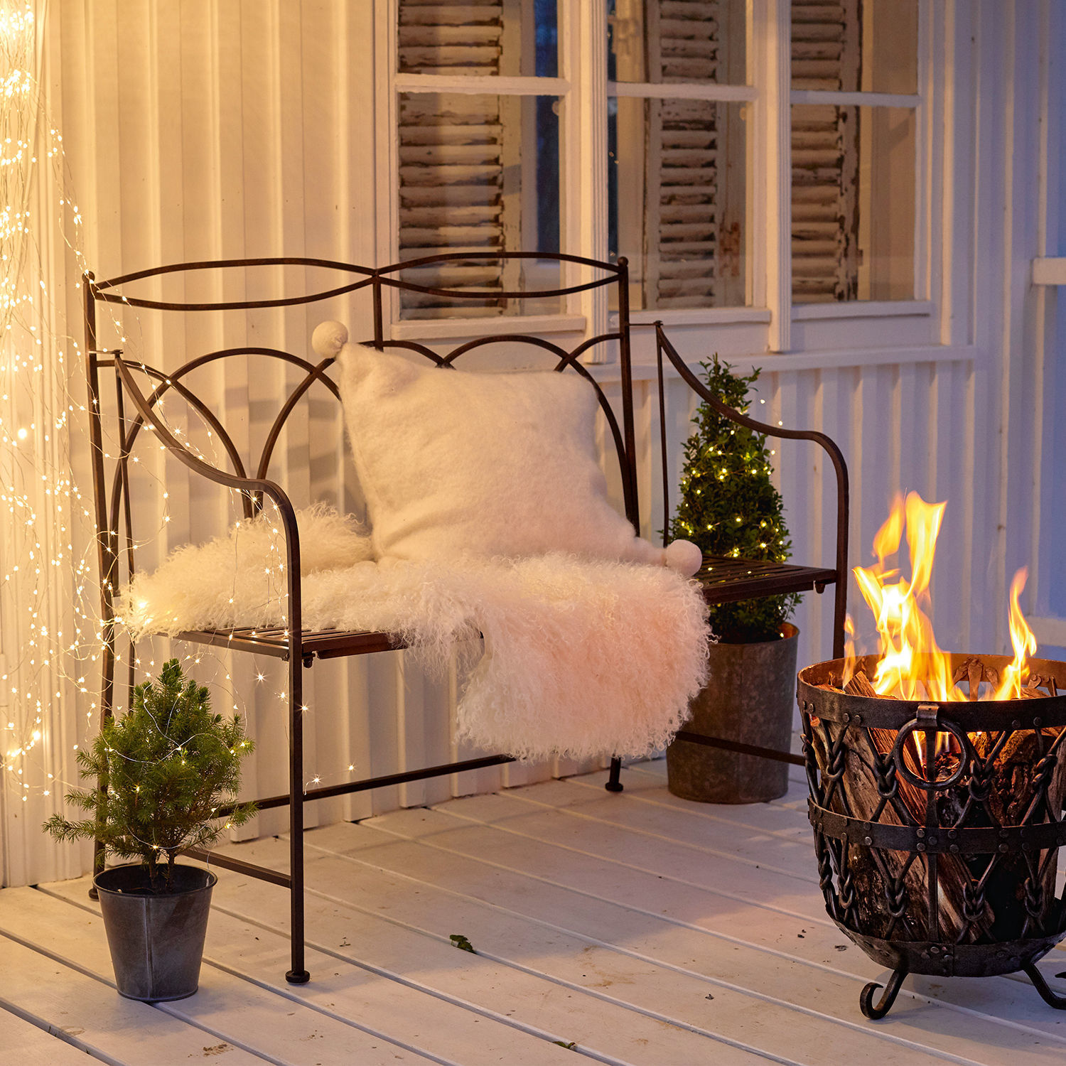 Bank Jablines Metallbank mit weißem Kissen und Fellüberwurf neben Feuerkorb auf Terrasse mit Lichterketten-Dekor