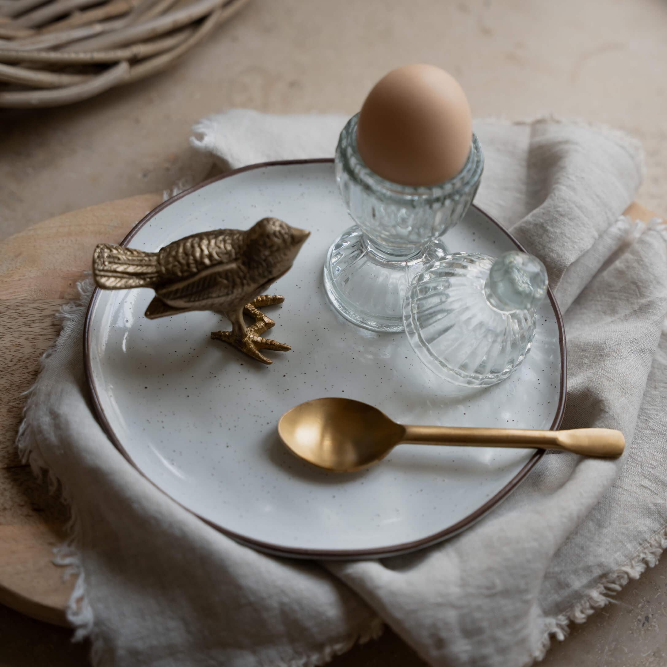 Glas-Eierbecher mit Deckel und braunem Ei auf weißem Teller mit Messinglöffel und Vogel-Figur