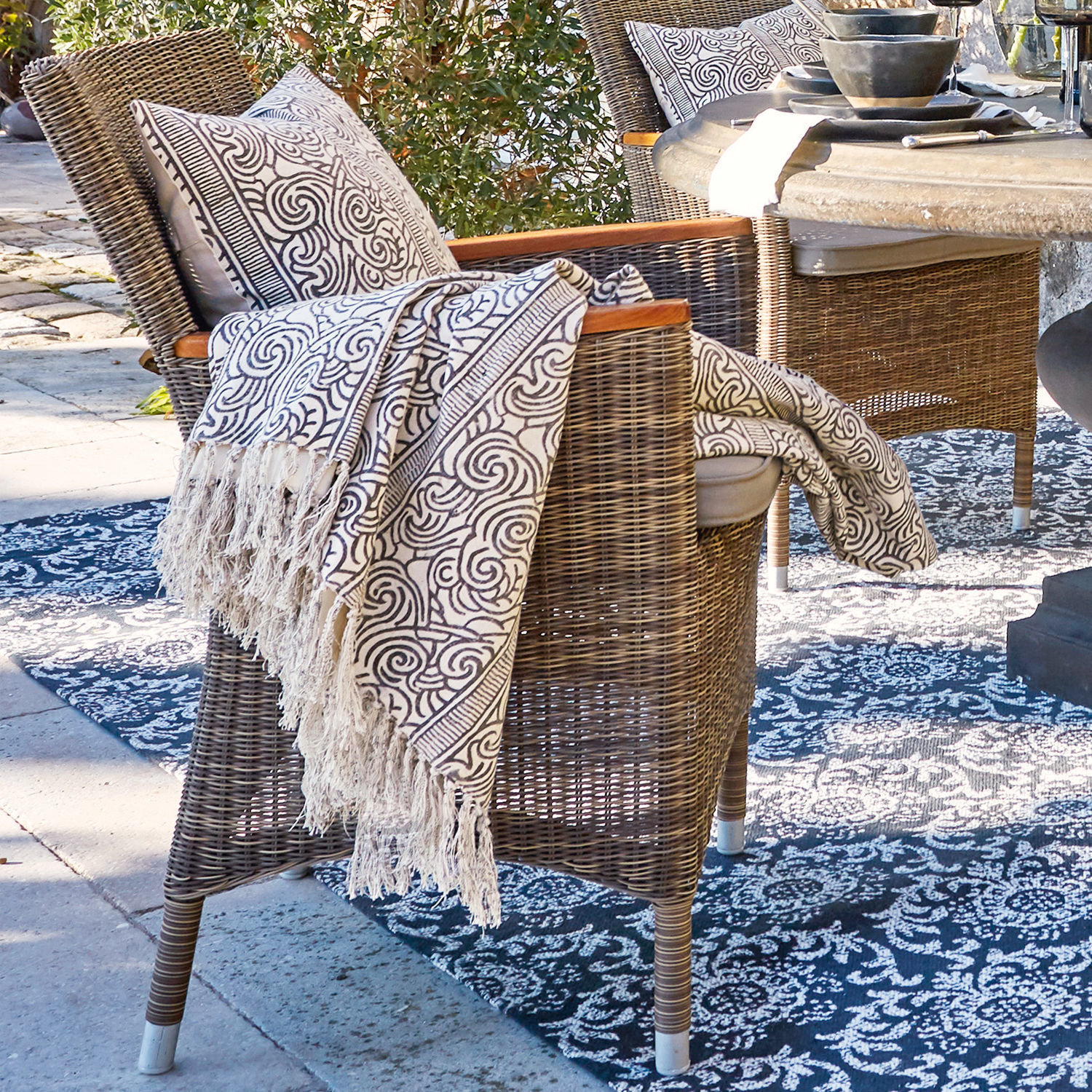 Brauner Rattanstuhl mit weißem Kissen und gemustertem Plaid auf Outdoor-Teppich im Garten