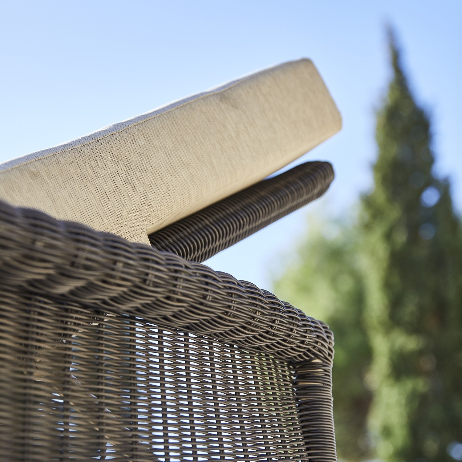Nahaufnahme eines braunen Rattanstuhls mit cremefarbenem Kissen vor blauem Himmel und Grün