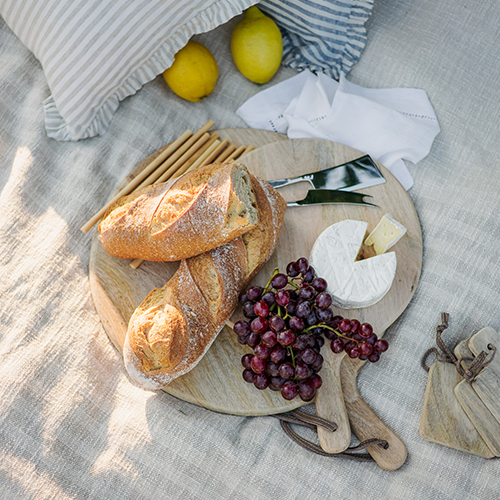 Runde Holzservierplatte mit Baguette, Camembert und roten Trauben bei Picknick auf Leinentuch