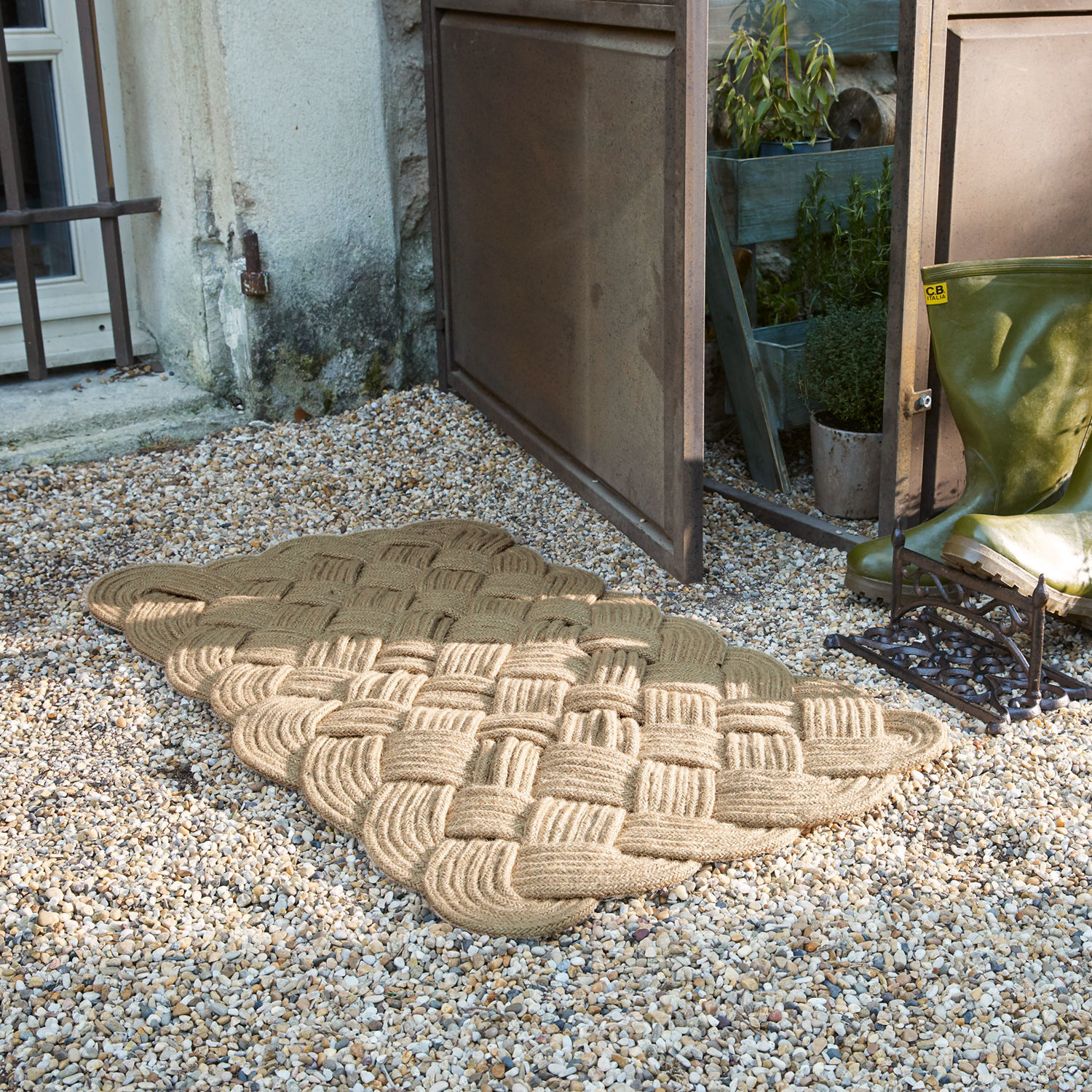 Beige Fußmatte aus geflochtenem Seil auf Kiesboden vor offener Tür neben grünen Gummistiefeln
