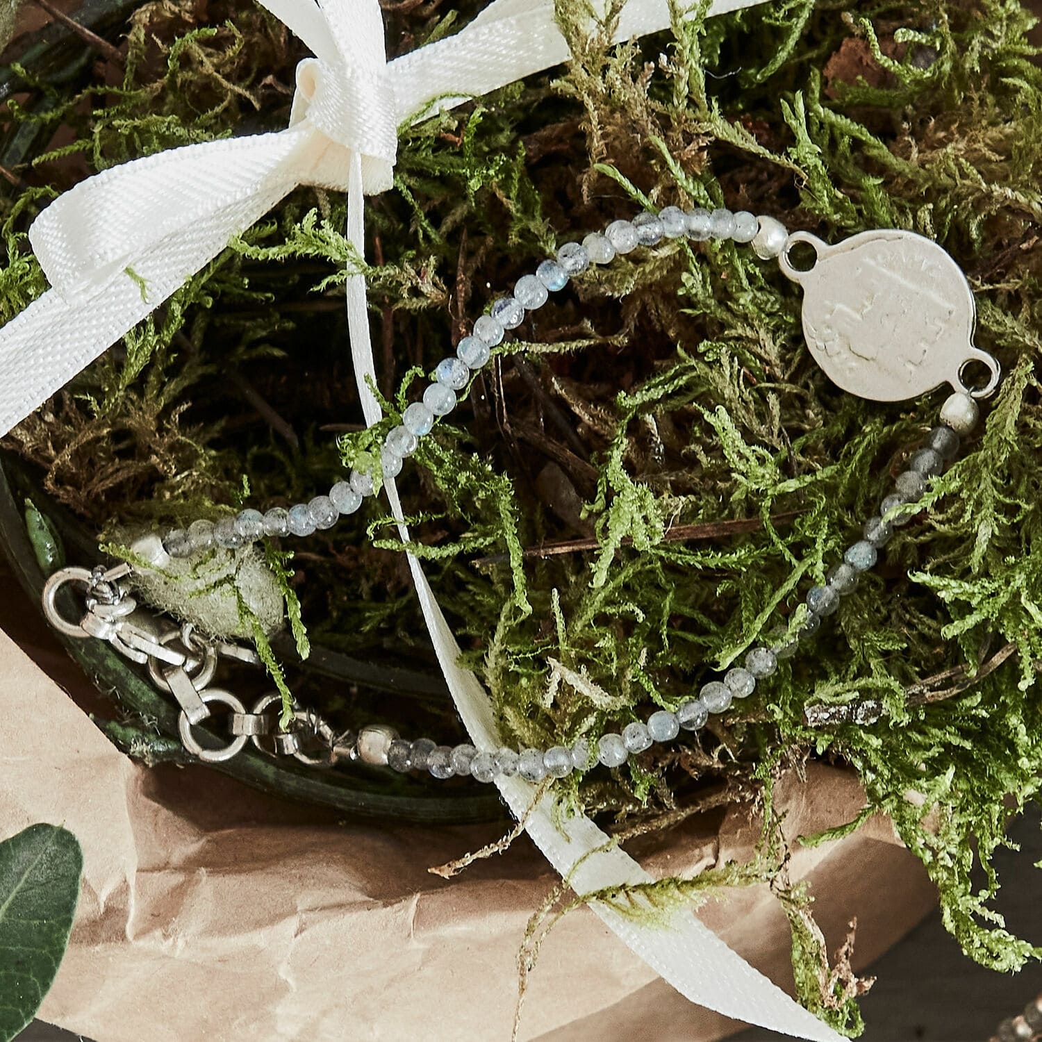 Feines Armband aus grauen Steinen mit Metallanhänger auf grünem Moos und weißem Band