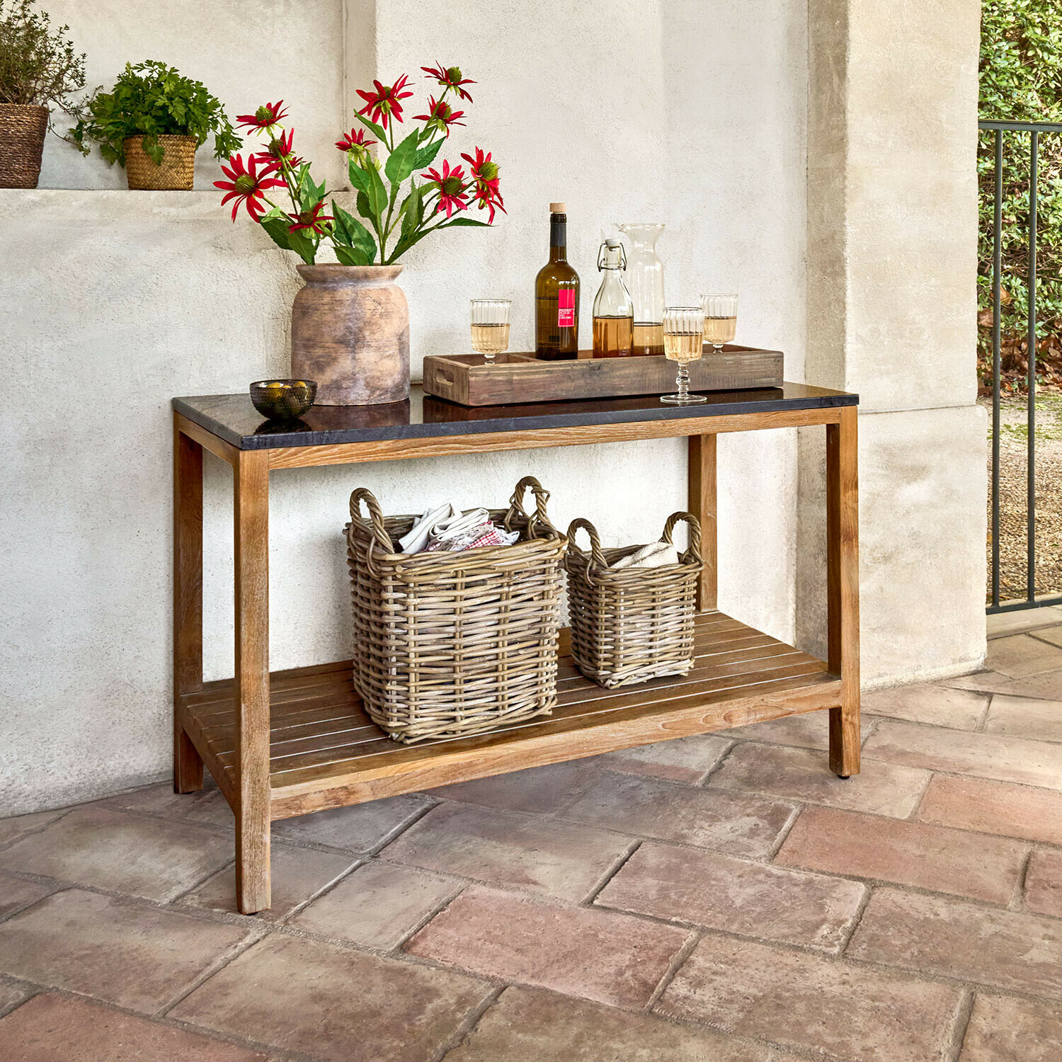 Outdoor-Küchentisch Corviano Konsole mit Holzrahmen und schwarzer Tischplatte, dekoriert mit Vase und roten Blumen sowie Weidenkörben darunter