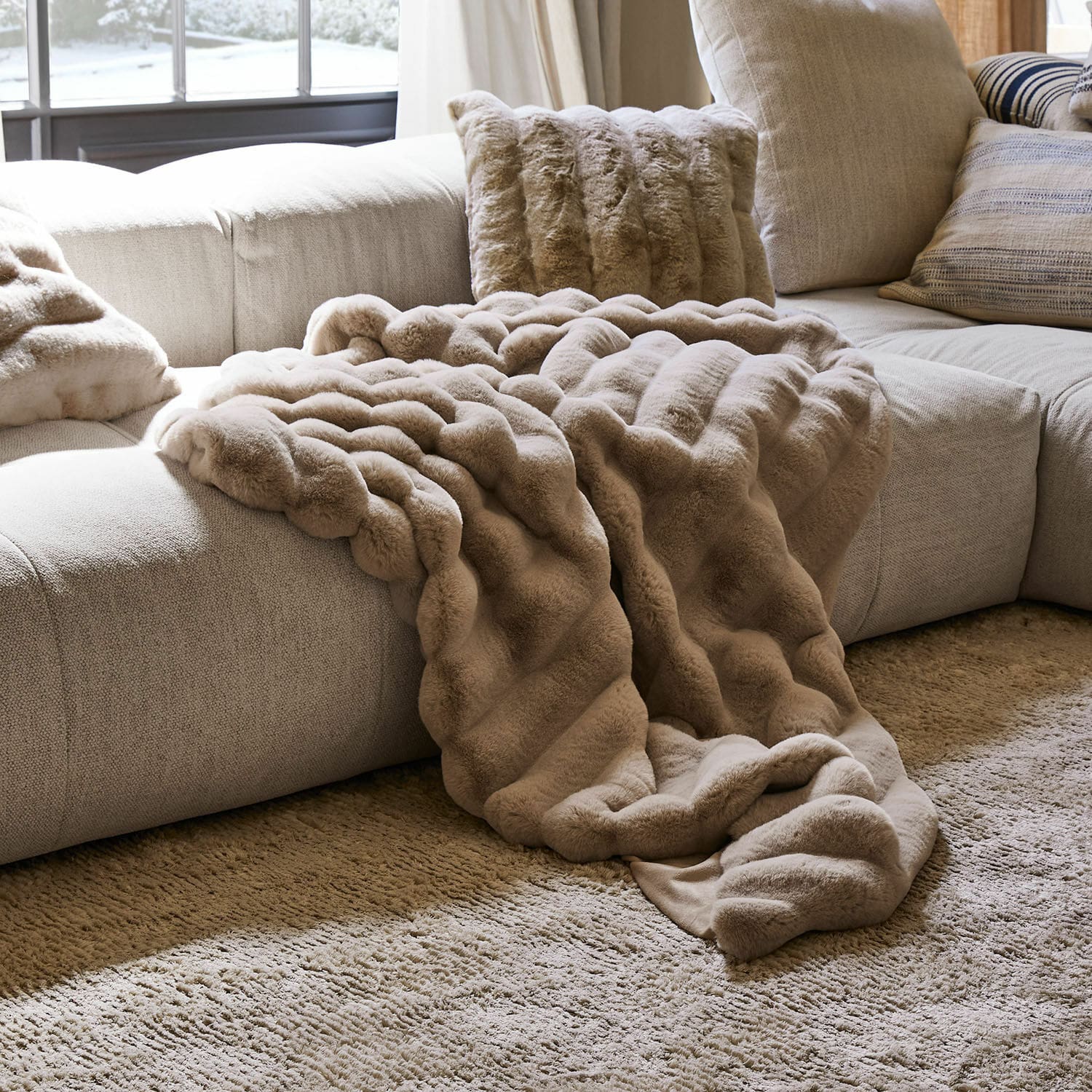 Plaid Caelira Beige weiche Felldecke und Kissen auf hellgrauem Sofa in gemütlichem Wohnzimmer