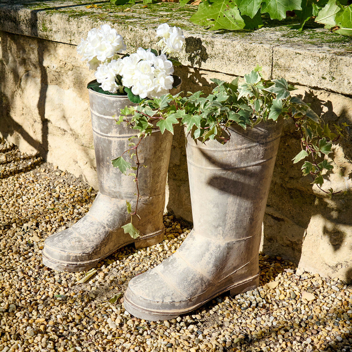 Alte graue Gummistiefel als Pflanzgefäße mit weißen Blumen und grünem Efeu im Garten