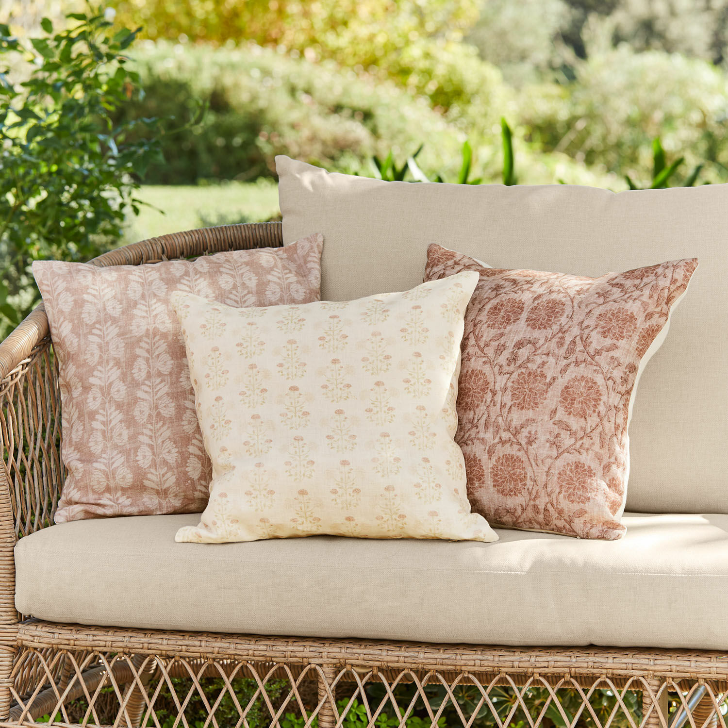 Kissenhülle 3er Set Tarouja Drei dekorative Baumwollkissen in Beige-Rosa mit floralen Mustern auf einem Rattansofa im Garten