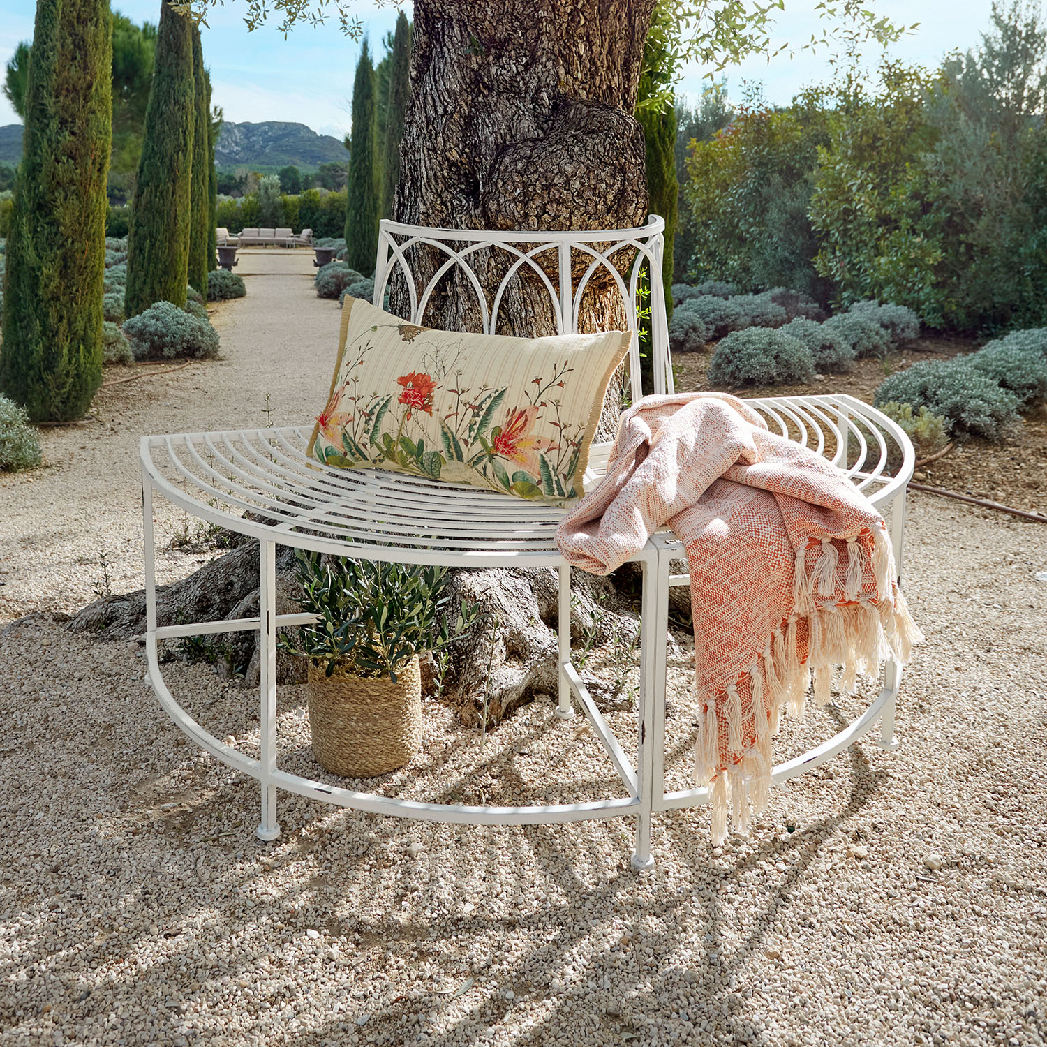 Weiße Metallbank um einen Baum mit floralem Kissen und rosa Decke auf Kiesweg im Garten