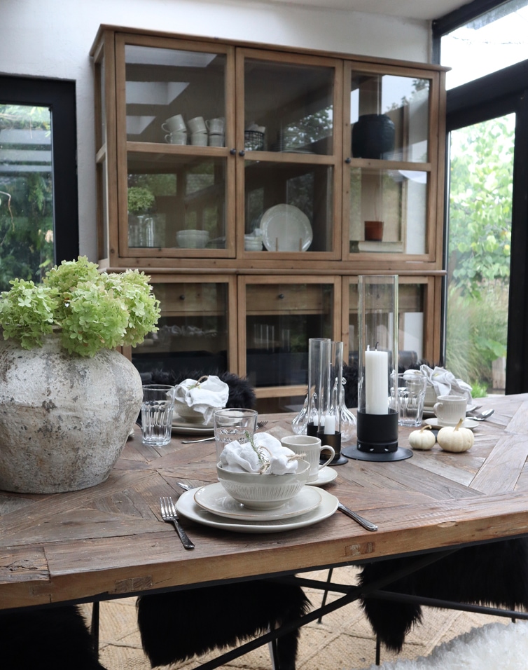 Rustikaler Holztisch mit Geschirr und Dekorationen in heller Orangerie mit Holz-Glas-Vitrine
