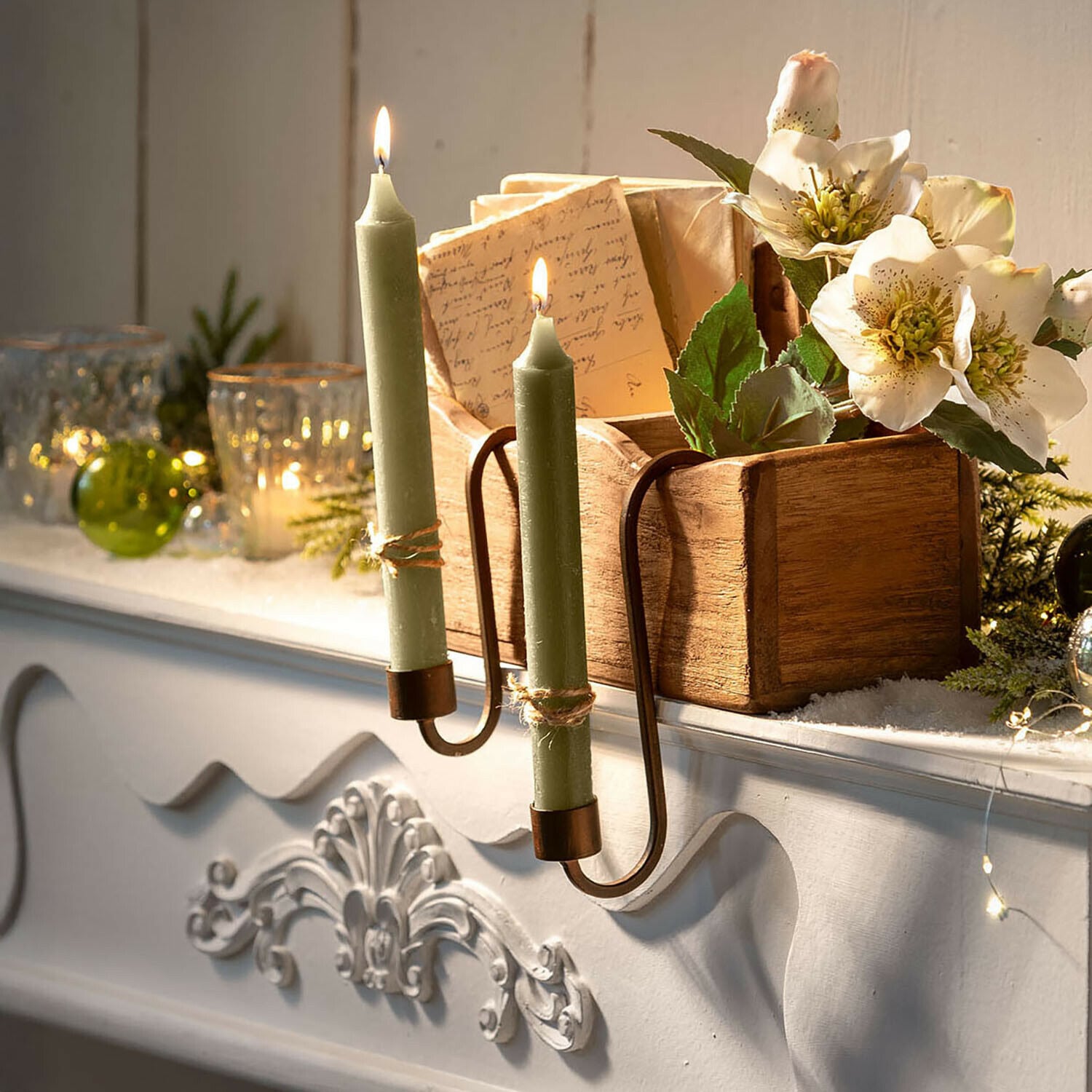 Grüne Kerzen im Metallkerzenhalter an weißem Kamin mit Holzkiste und Blumen dekoriert