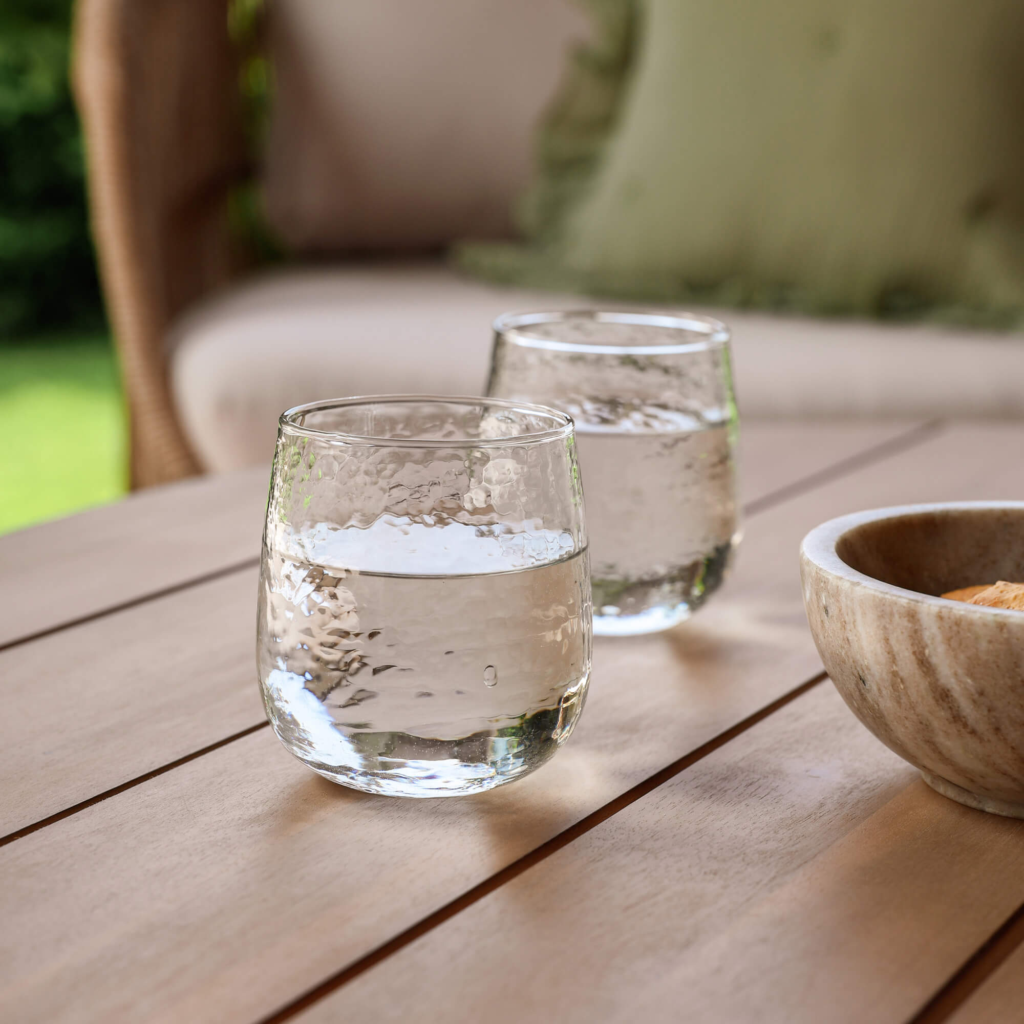 Zwei klare Wassergläser auf einem Holztisch im Garten neben einer Steinschale
