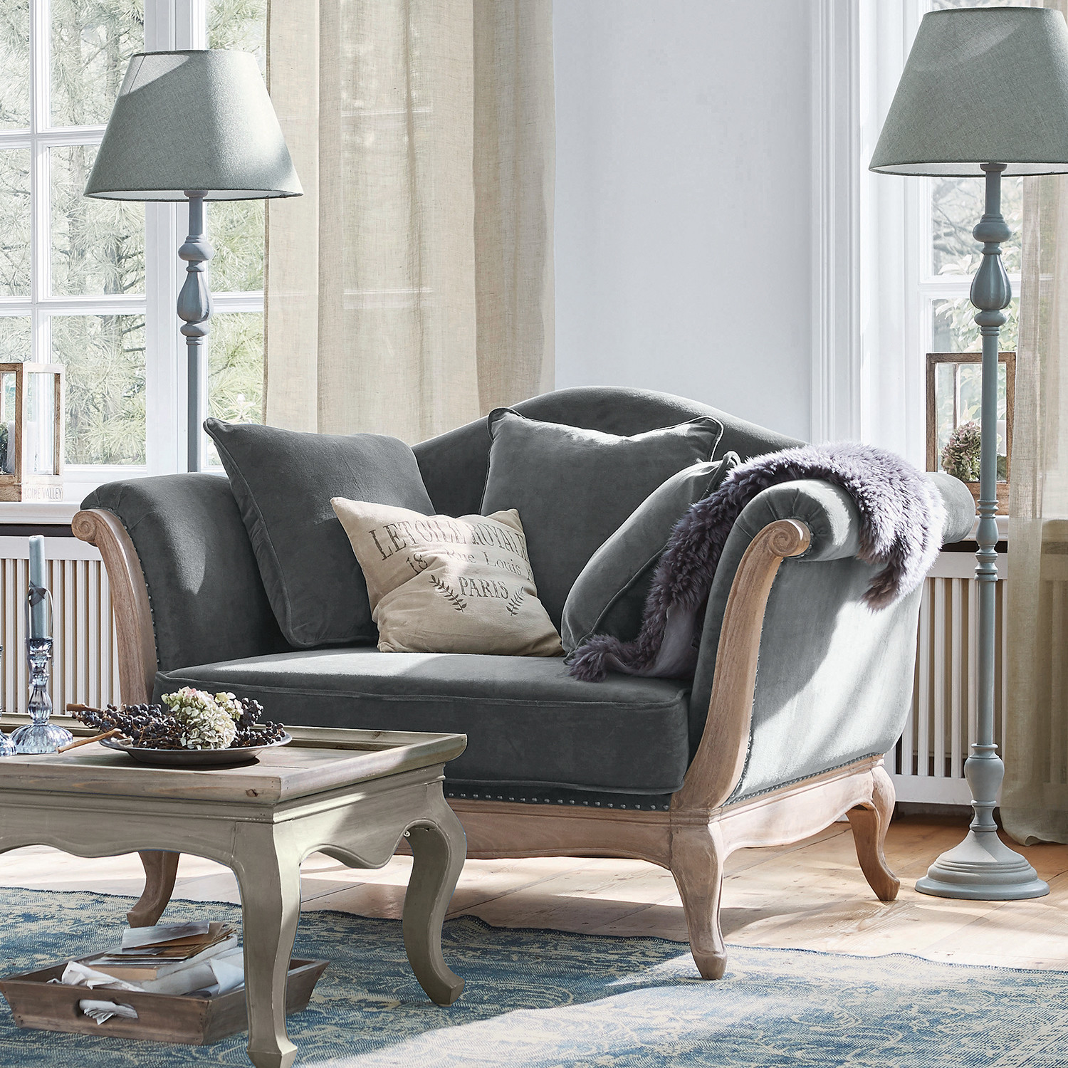 Sessel Sumerset Graues Samtsofa mit Holzrahmen und Kissen in hellem Wohnzimmer mit Stehlampen