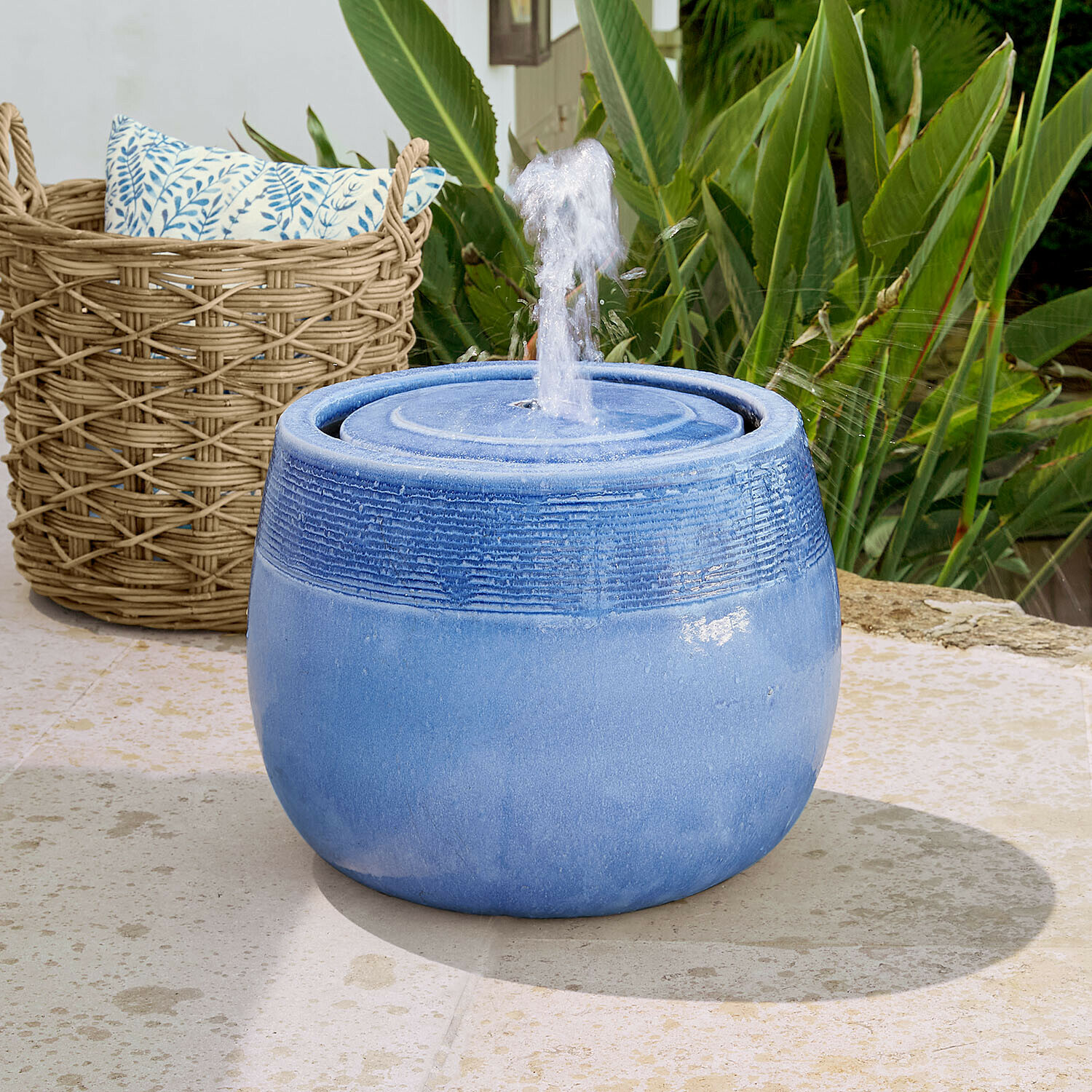 Brunnen Seravia Blaue Keramik-Gartenbrunnen mit spritzendem Wasser vor grünen Pflanzen und Weidenkorb