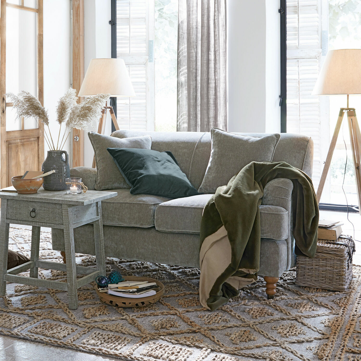 Graues Sofa mit weichen Kissen und grünem Überwurf in hellem Wohnzimmer mit Holzlampen und Teppich