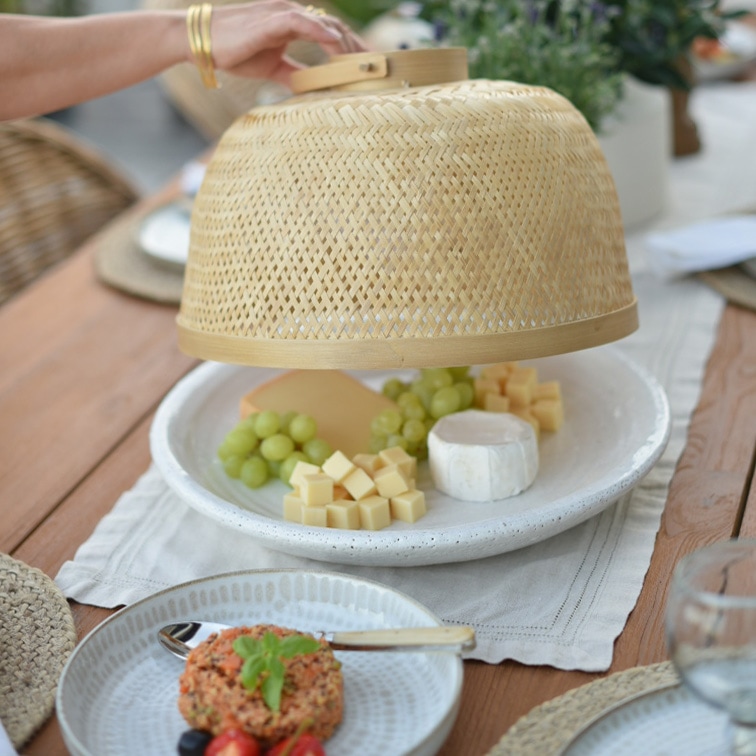Natürliche Bambus-Abdeckhaube schützt Käse und grüne Trauben auf weißem Teller auf Holztisch