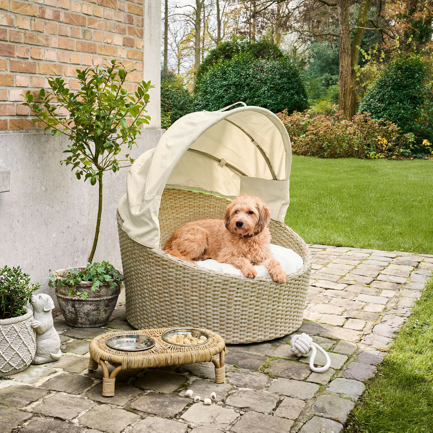 In- & Outdoor-Haustierkorb Brinelza Rundes Hundebett aus beigem Rattan mit cremefarbenem Dach und Kissen auf gepflasterter Terrasse