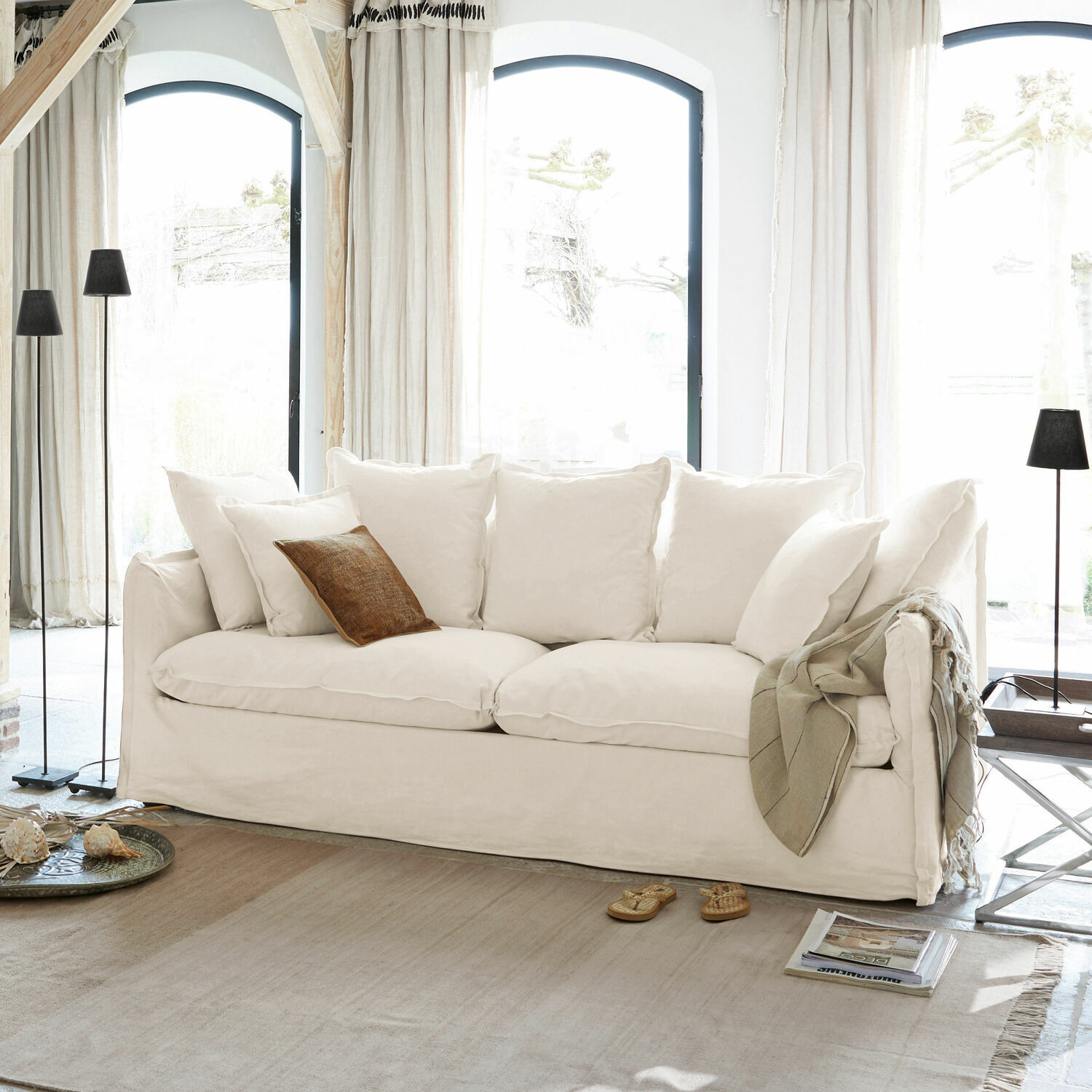 Sofa Tienen Weißes Sofa mit weichen Kissen und braunem Dekokissen in hellem Wohnzimmer mit großen Fenstern