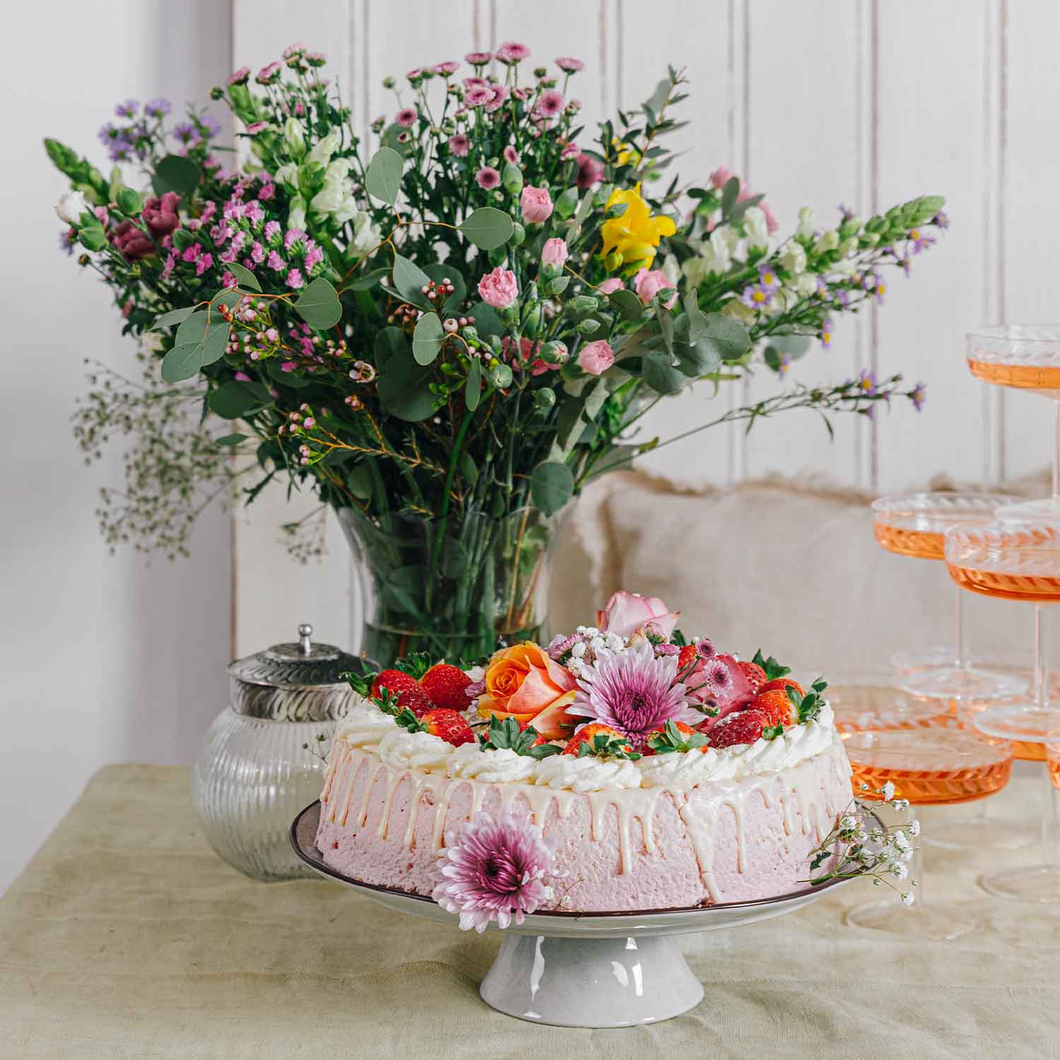 Rosafarbene Sahnetorte mit frischen Erdbeeren und Blumen auf weißem Tortenständer vor Blumenstrauß