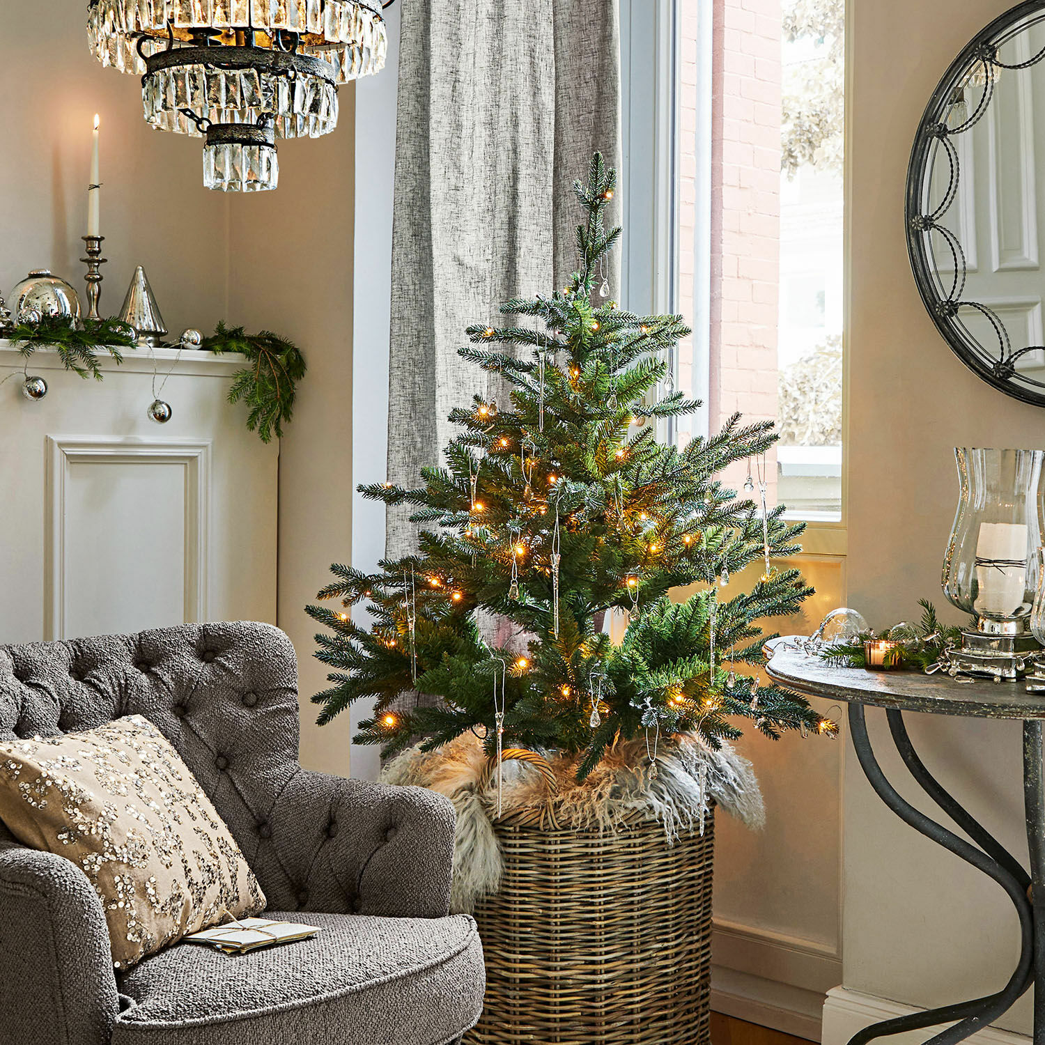 Kleiner Weihnachtsbaum im Korb mit LED-Lichterkette im gemütlichen Wohnzimmer mit grauem Sofa und Dekorationen