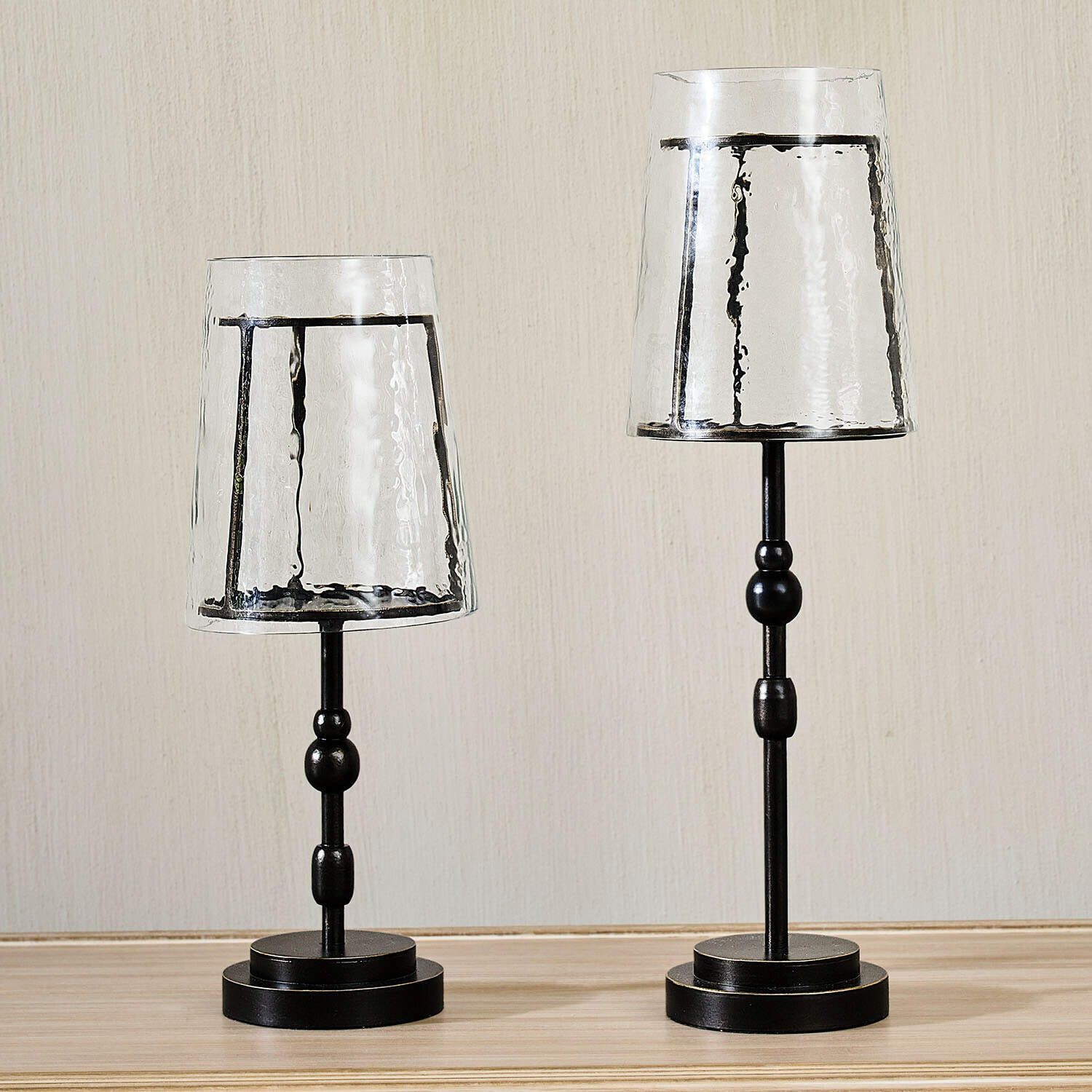 Zwei schwarze Metallkerzenhalter mit transparenten, strukturierten Glasschirmen auf Holzuntergrund