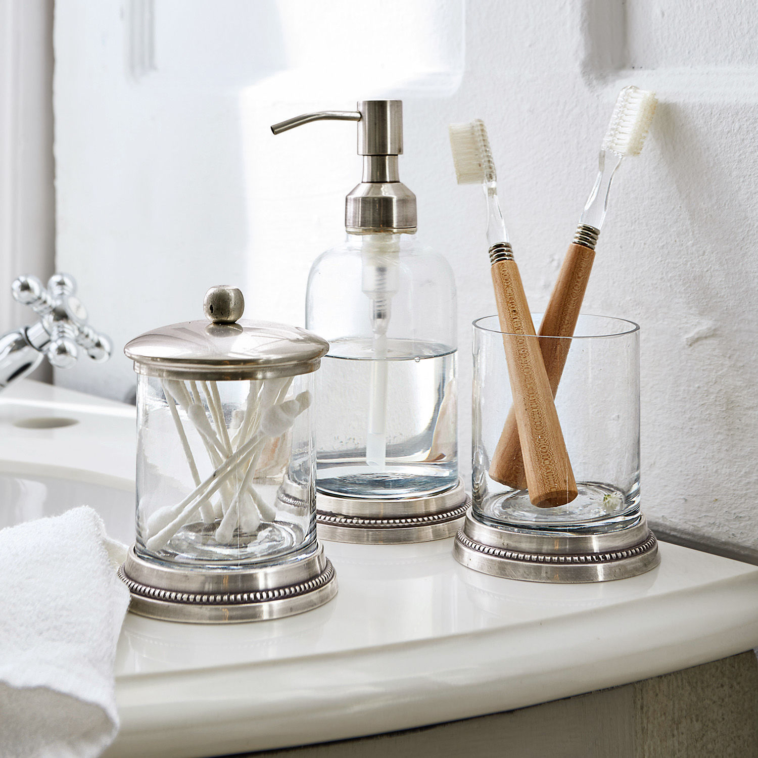 Glas-Badezimmeraccessoires mit Metallakzenten auf weißem Waschbecken mit Bambuszahnbürsten