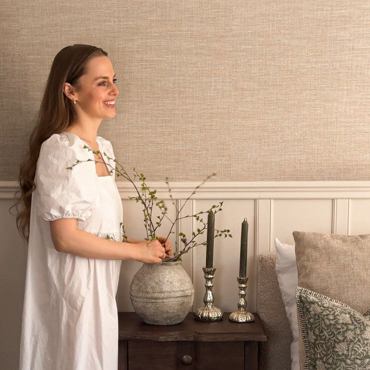 Frau im weißen Kleid arrangiert Zweige in grauer Keramikvase auf Holztisch mit Kerzen