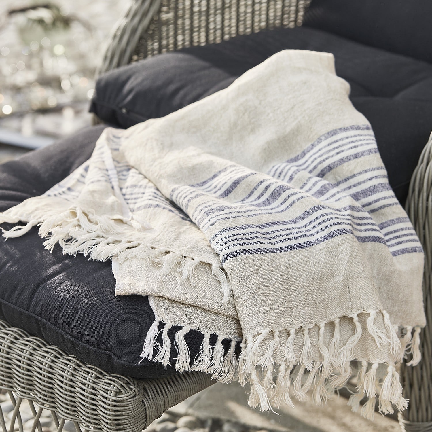 Beige Leinen-Decke mit blauen Streifen und Fransen auf schwarzem Kissen eines Rattansessels