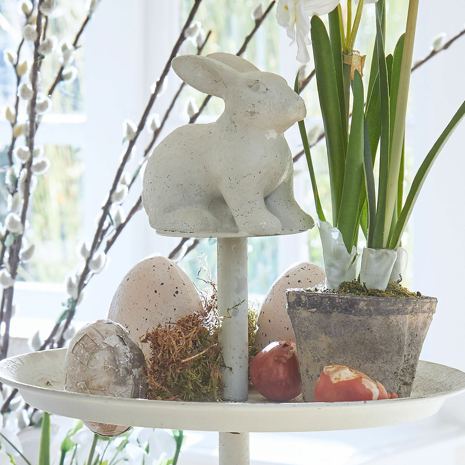 Dekorativer weißer Keramik-Hase auf Ständer mit Blumentopf und pastellfarbenen Deko-Eiern