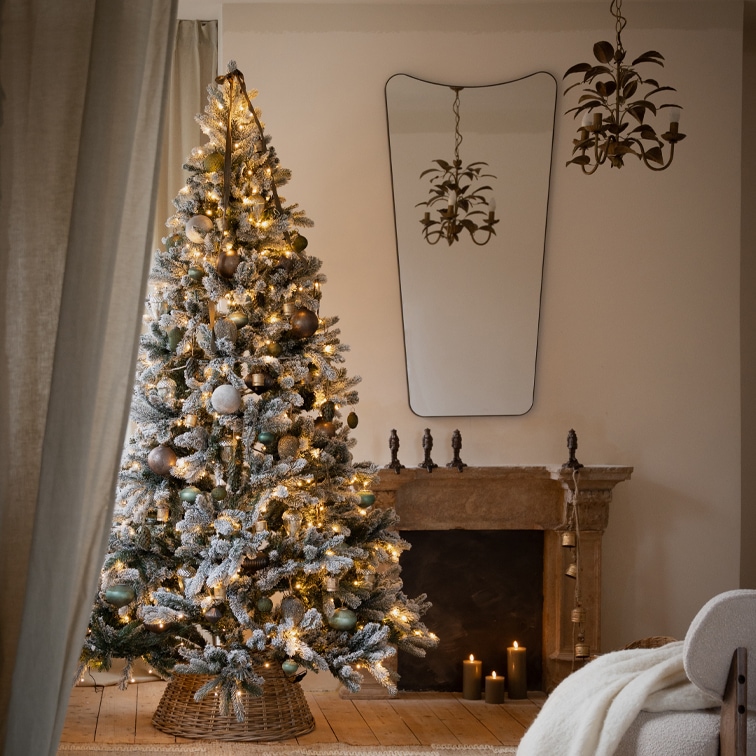 Oberflockierter Weihnachtsbaum mit Lichterkette und Kugeln im Korbständer vor Kamin im Wohnzimmer