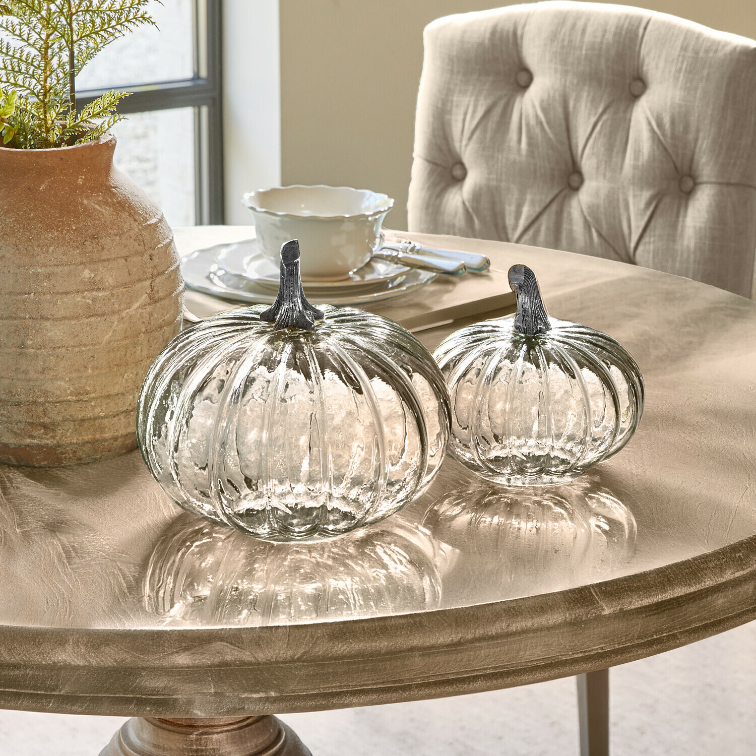Zwei transparente Glas-Kürbisse mit Metallstielen auf einem beigen Dekotisch