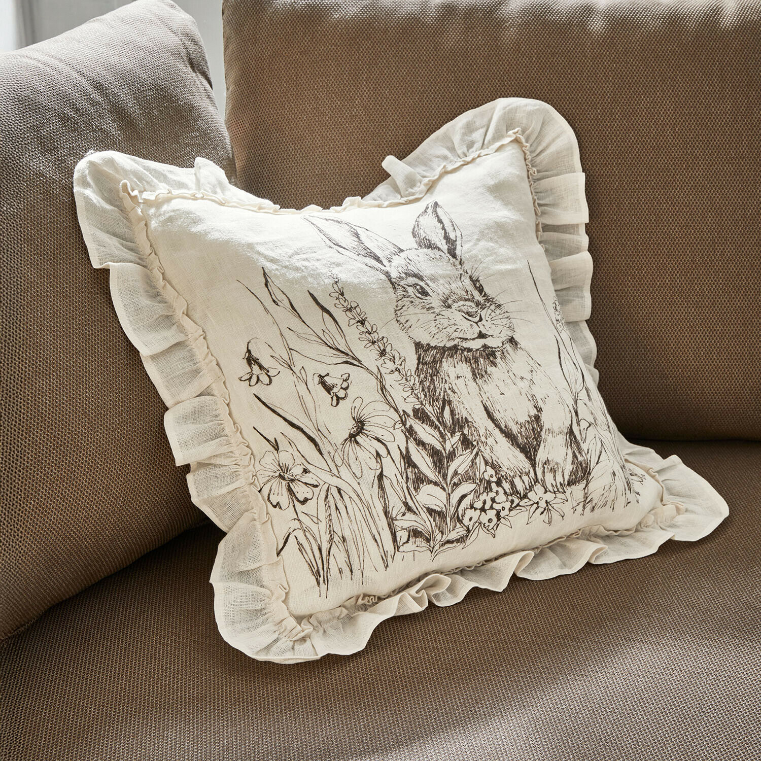 Kissenhülle Brionelle Beiges Leinenkissen mit Rüschen und Hasenmotiv mit Blumen auf braunem Sofa