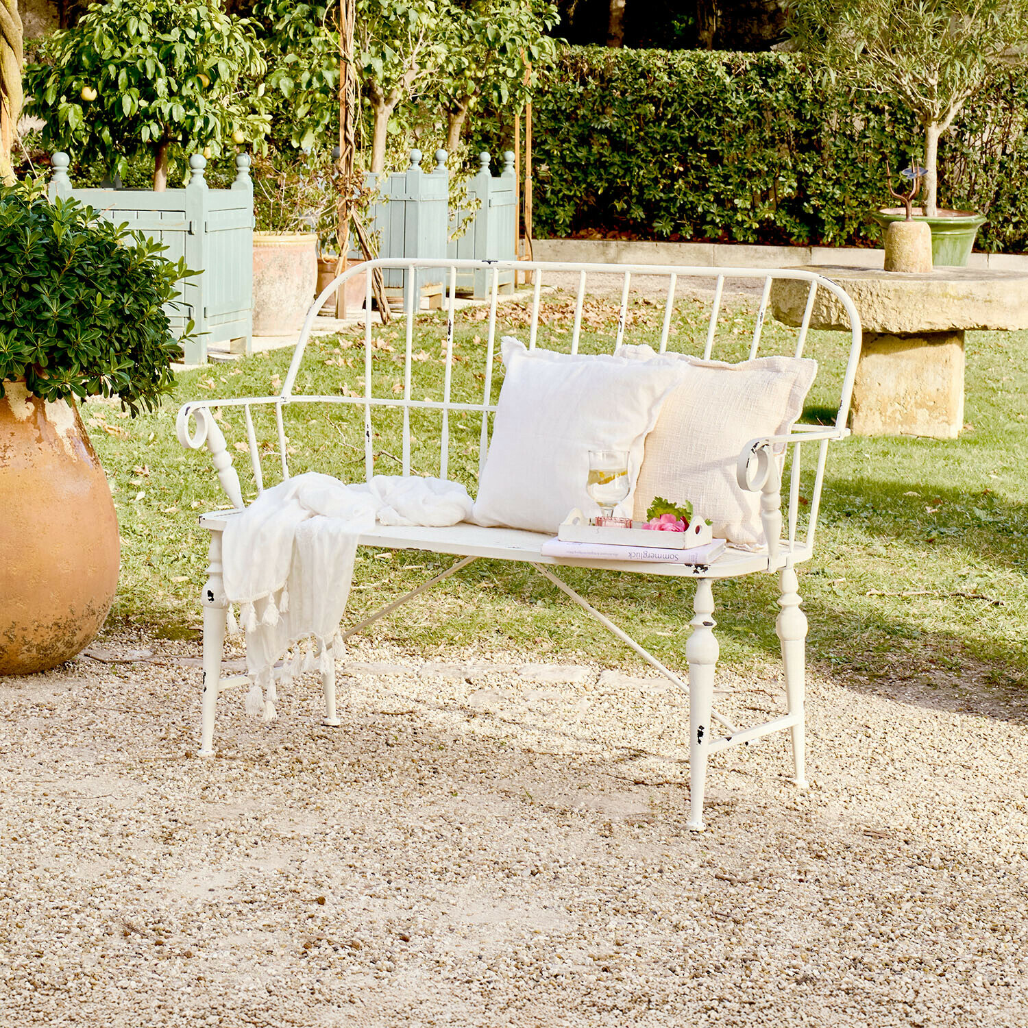 Weiße Metall-Gartenbank mit Kissen und Decke auf Kiesplatz in grünem Garten