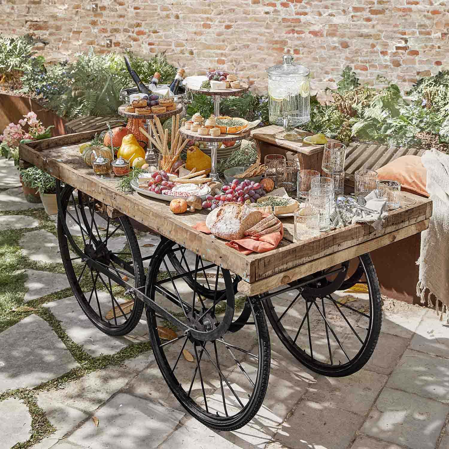 Rustikaler Holzwagen mit schwarzen Rädern, bestückt mit Snacks und Getränken im Garten