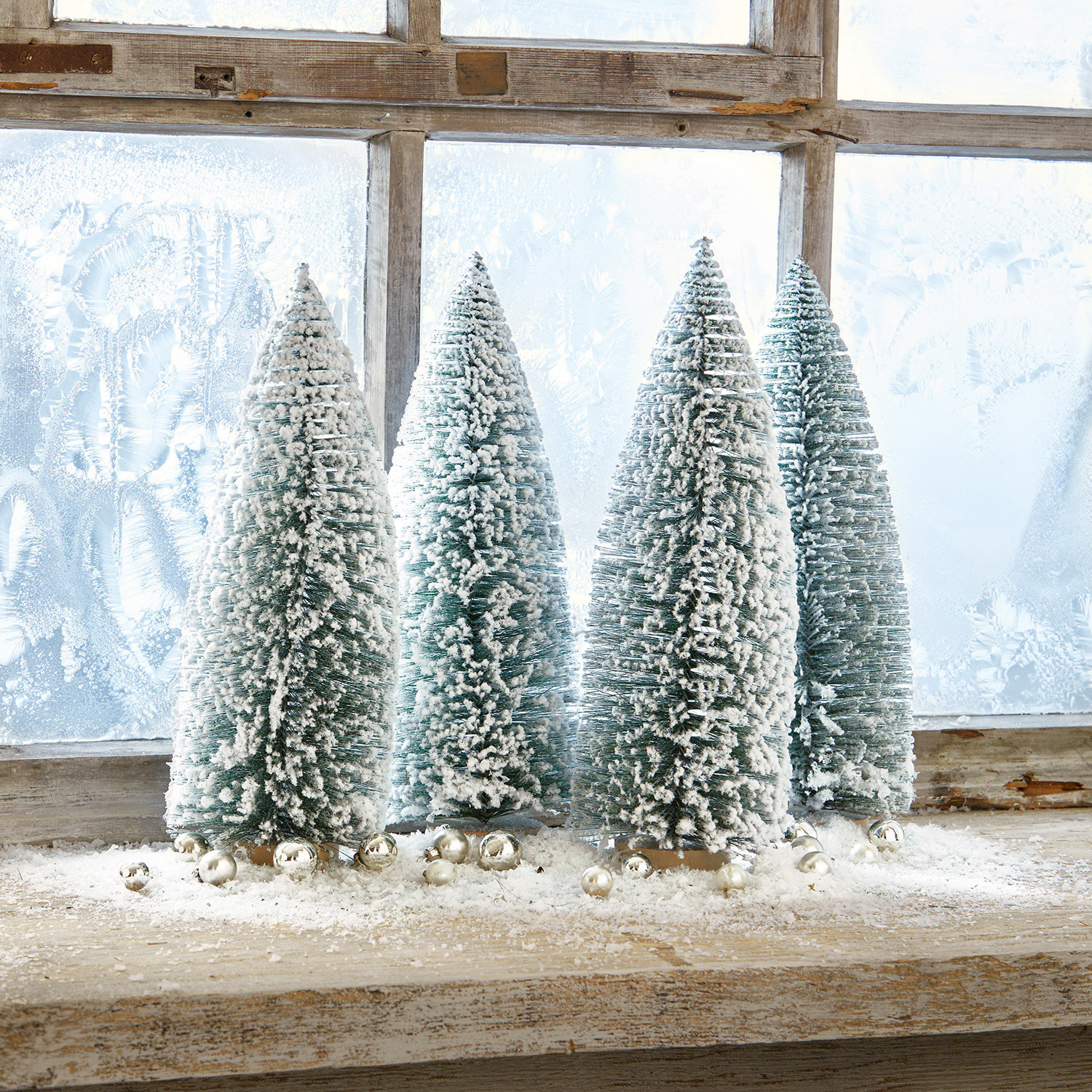 Deko-Baum 4er Set Landscape Vier grüne Kunststoff-Tannenbäume in Kegelform mit weißem Schnee auf einem Holzfensterbrett mit silbernen Kugeln
