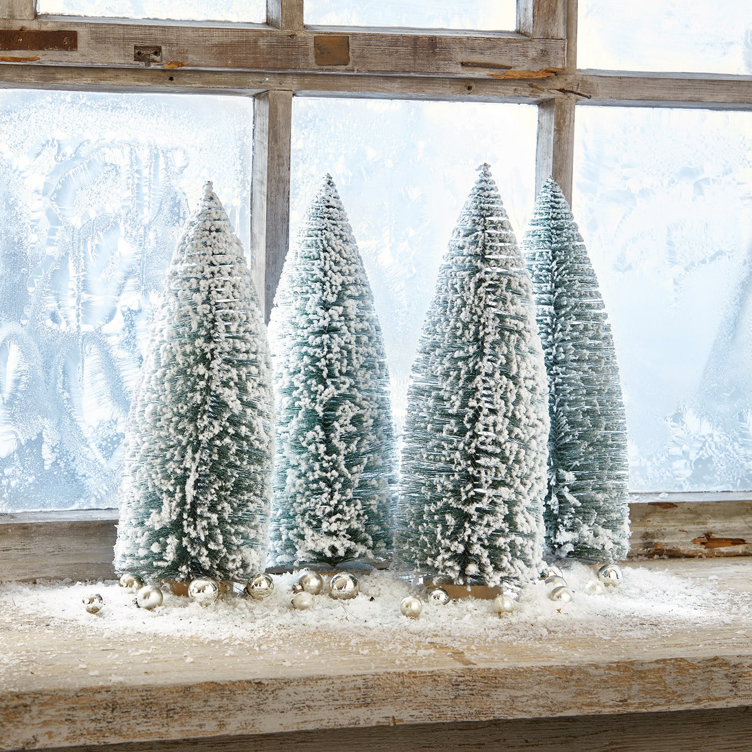 Deko-Baum 4er Set Landscape Vier grüne Kunststoff-Tannenbäume in Kegelform mit weißem Schnee auf einem Holzfensterbrett mit silbernen Kugeln